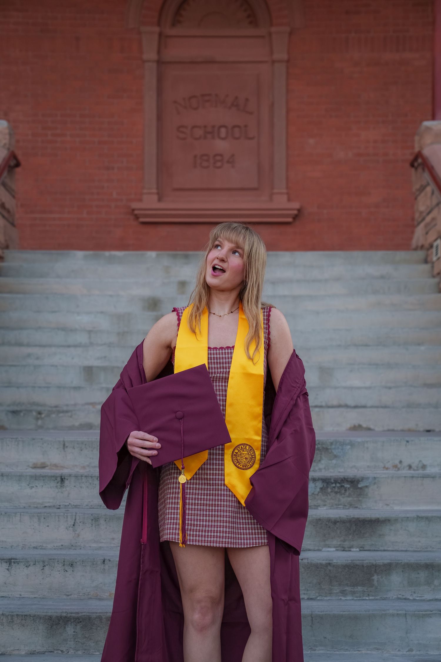 A graduate in maroon cap and gown with yellow stole poses on stone steps for a series of celebratory photos.