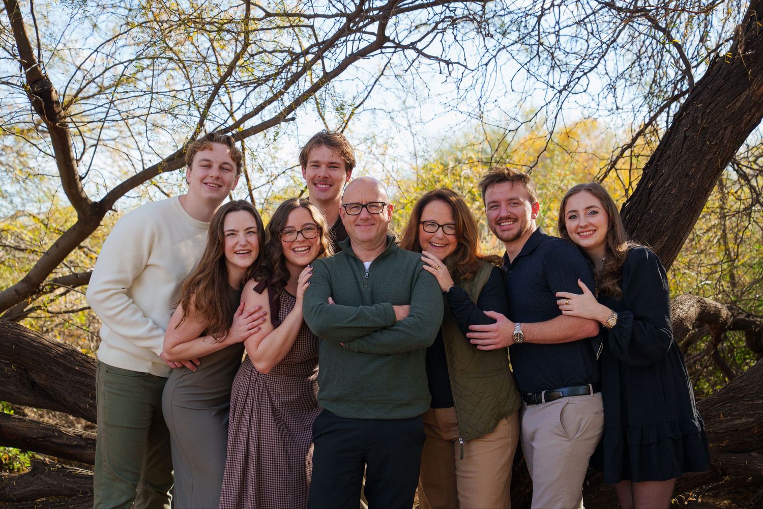 A joyful family of eight posing together outdoors among autumn trees in a warm group portrait.
