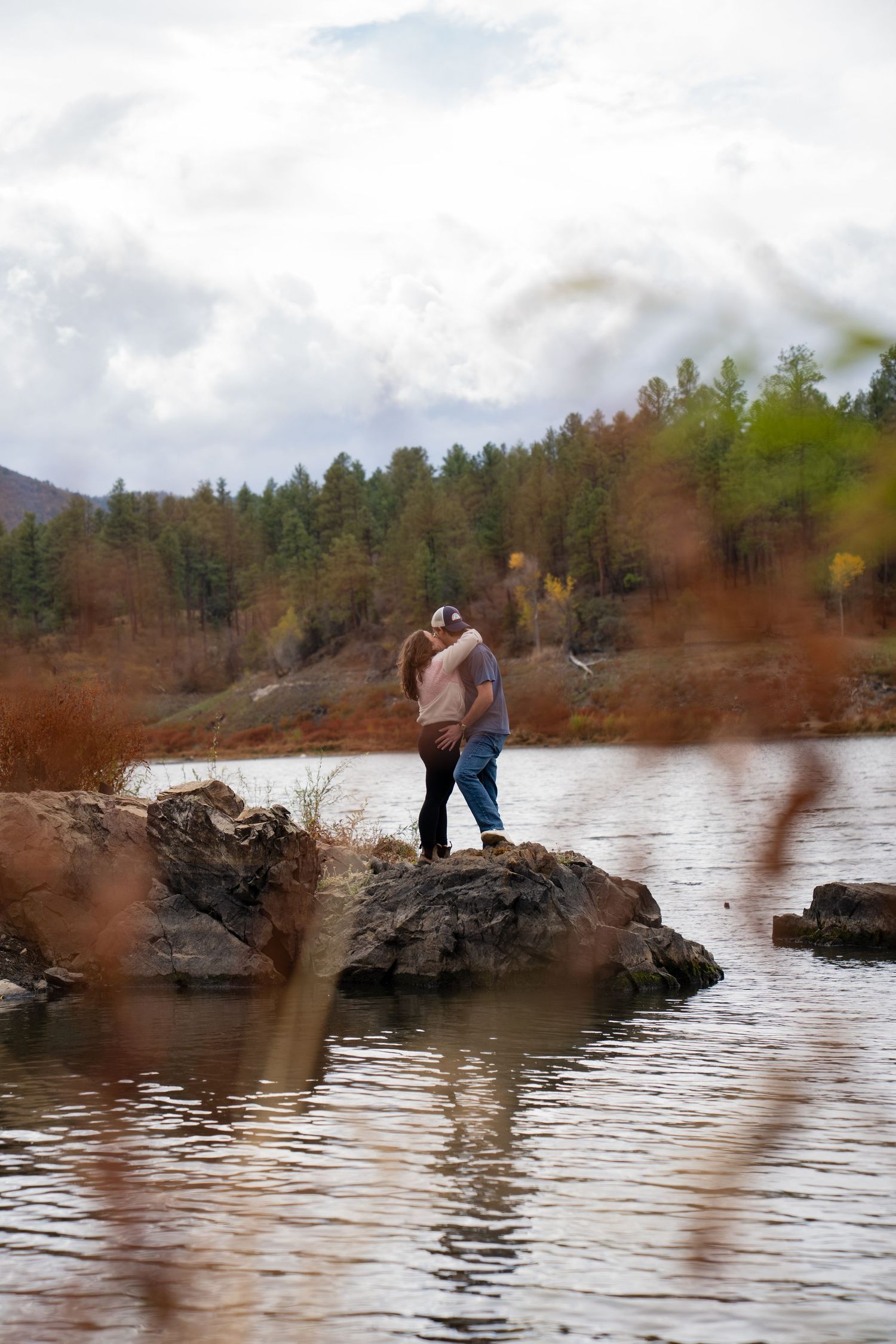Couple stands on rocky shore beside lake with pine forest in background.