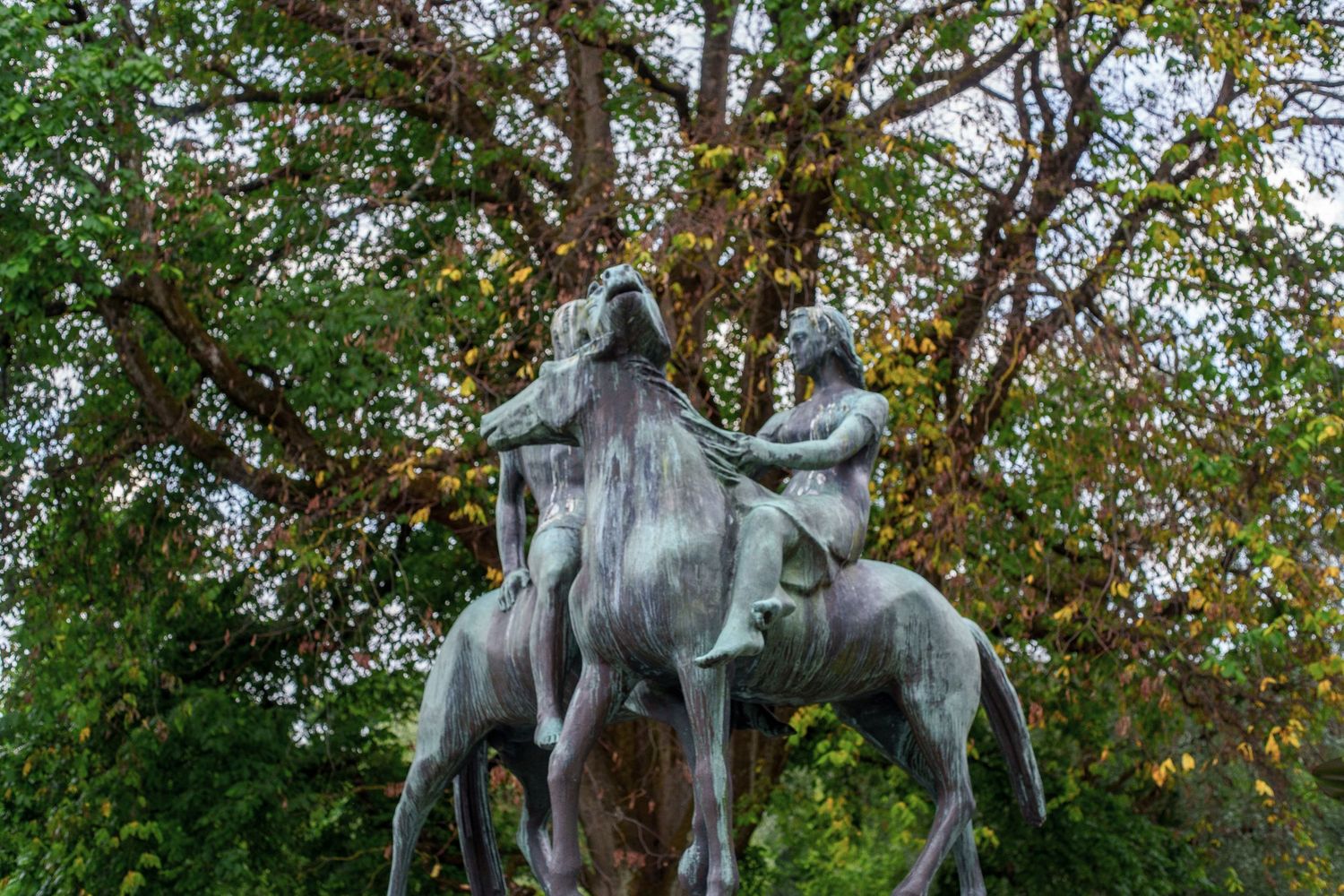Bronze statues of dancing figures arranged in a circle under green trees.
