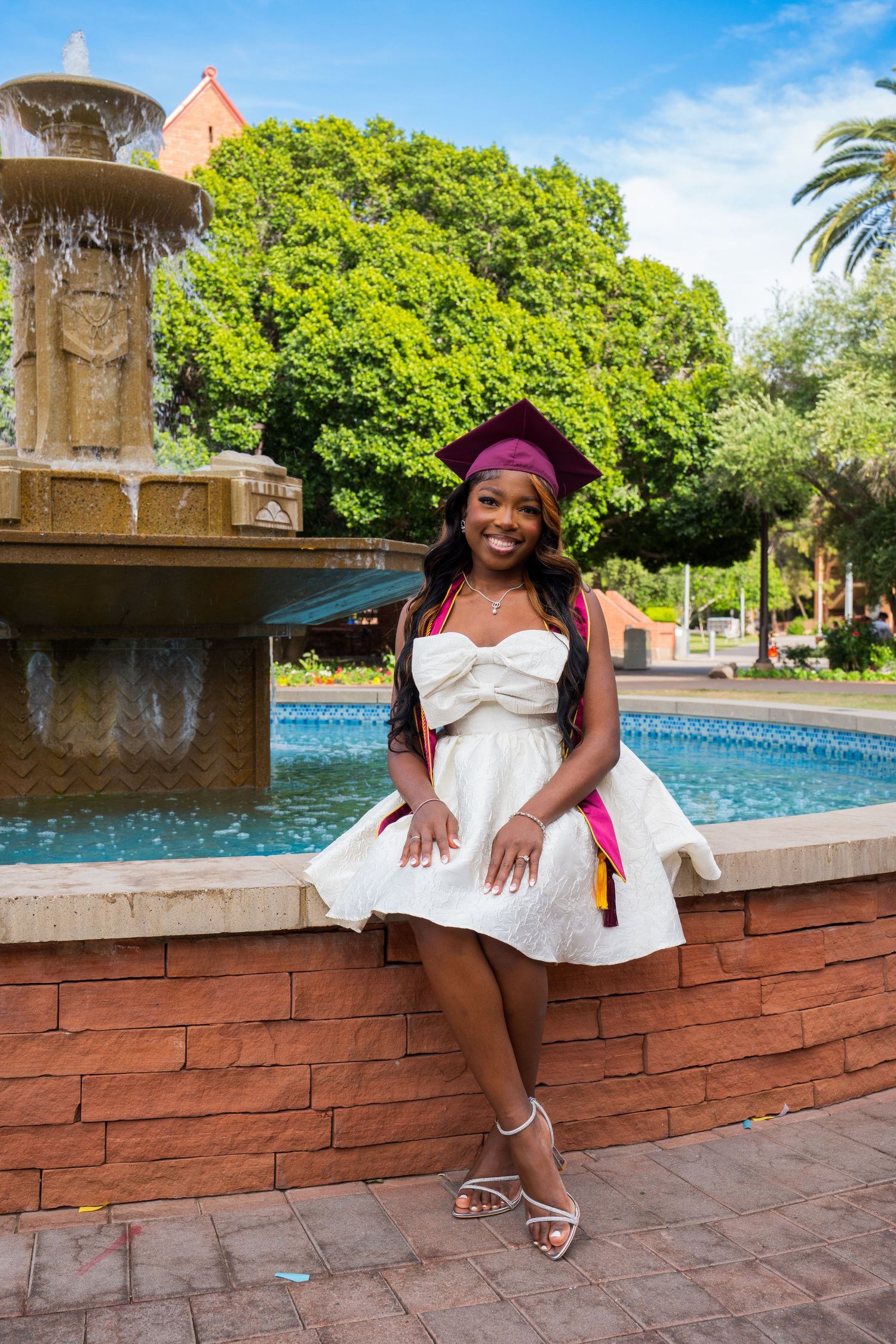 Person in white dress and burgundy hat sits on brick ledge near fountain in sunny outdoor setting.