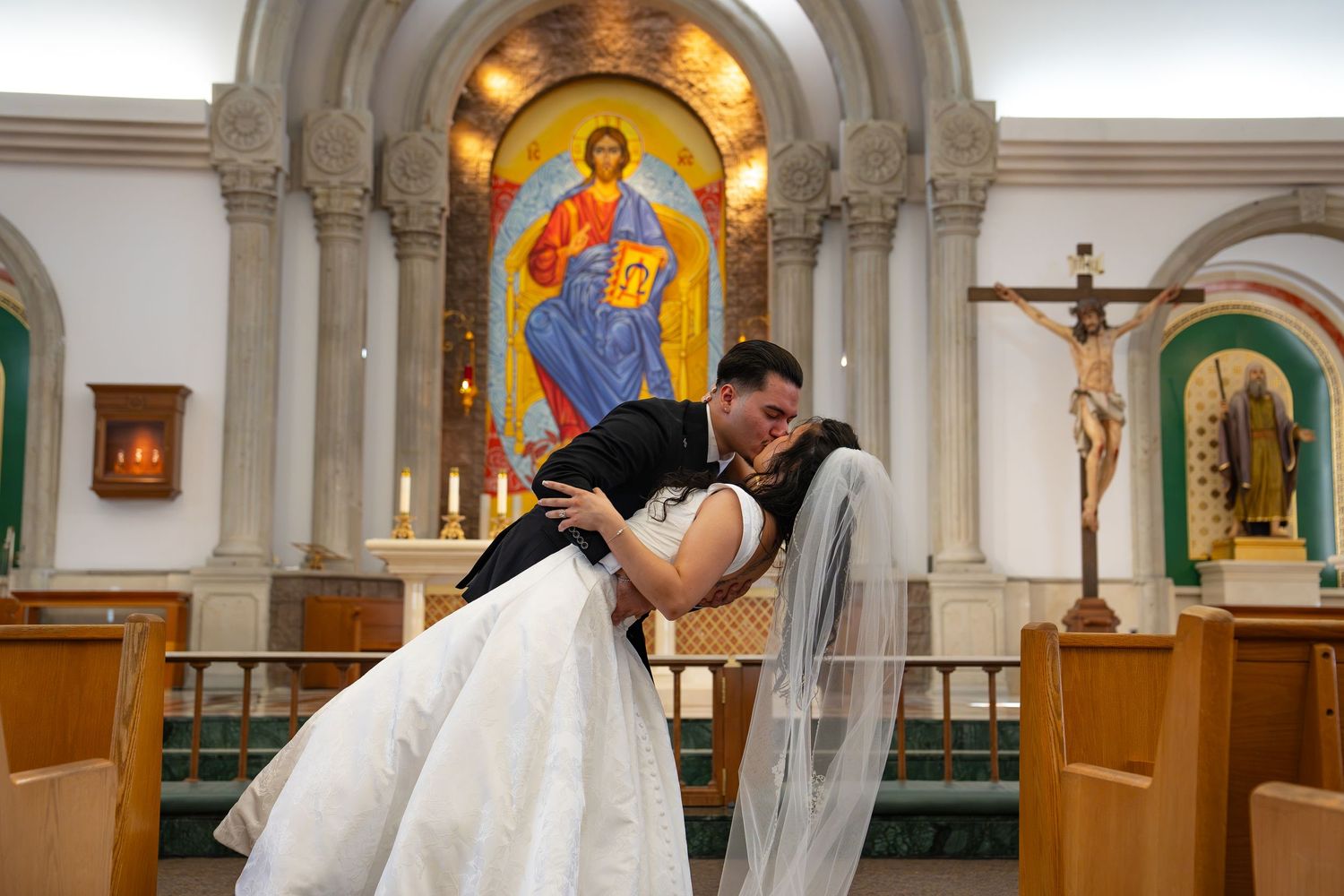 Bride and groom share a romantic dip kiss at church altar with Byzantine icon and crucifix in background.
