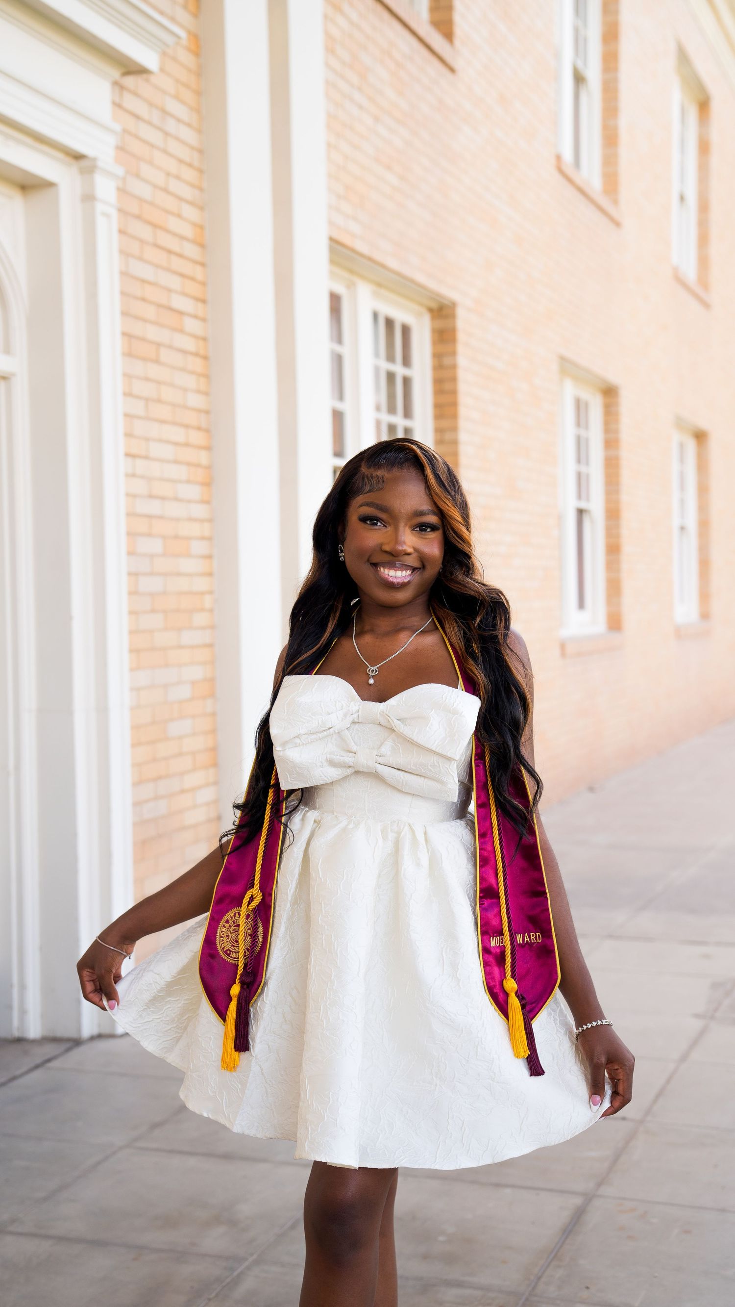 Graduate in white dress and pink stole poses joyfully against brick building in series of celebratory shots.