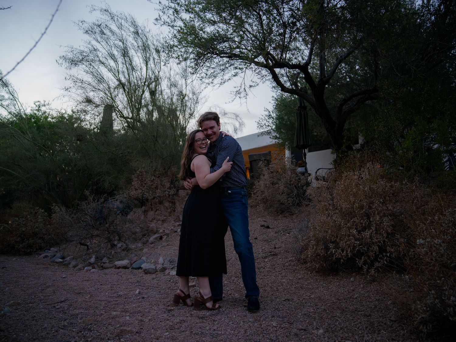 Couple embracing outdoors at dusk near desert trees and shrubs, woman in black dress and man in jeans.