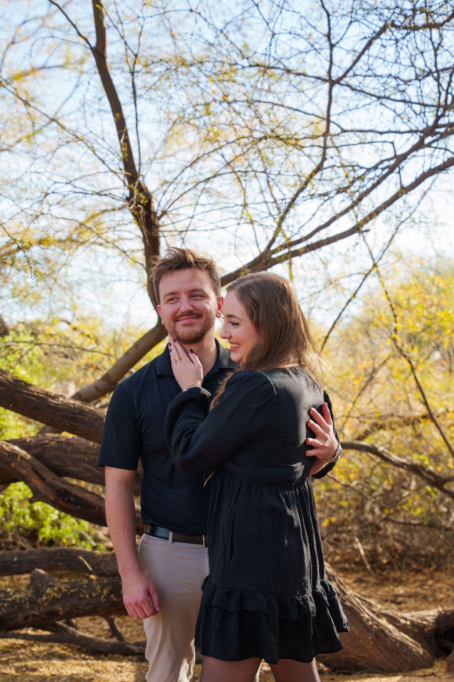 A couple embraces and smiles outdoors among bare trees, both dressed in black clothing on a sunny day.