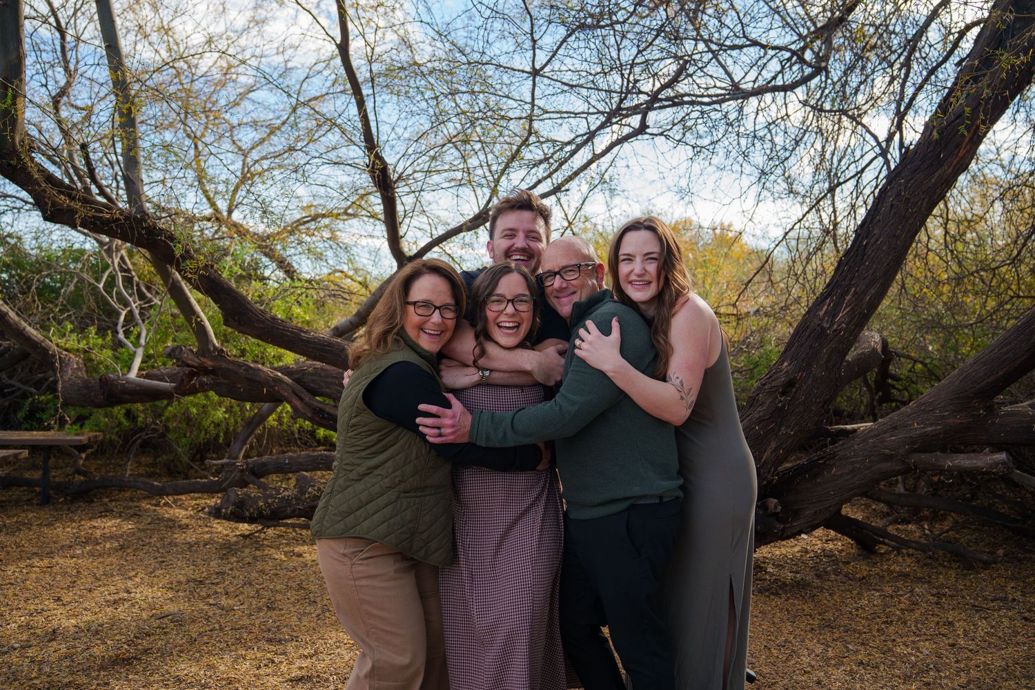 Five friends smiling and hugging outdoors among bare trees on a sunny day.