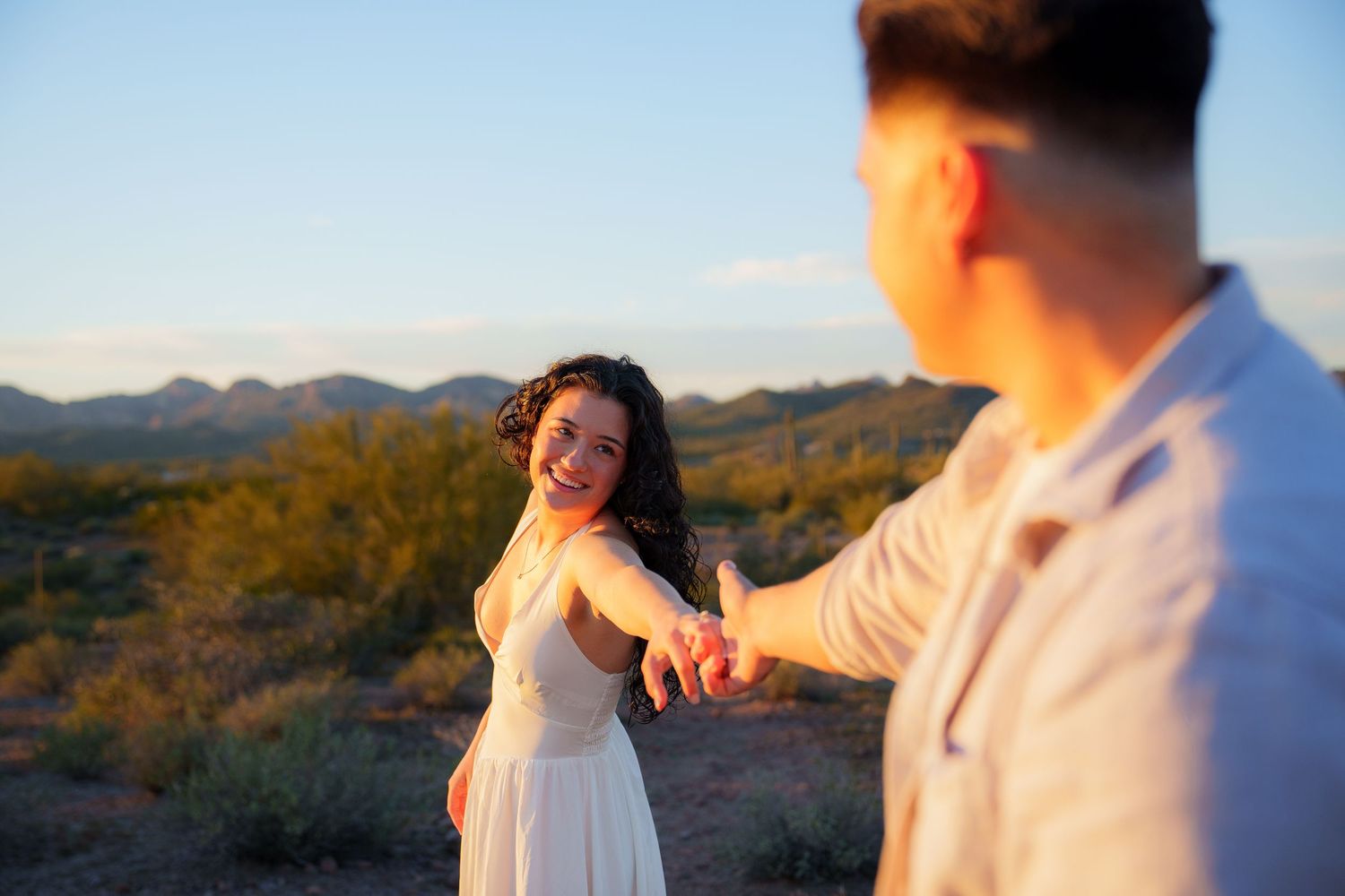 A couple holds hands at sunset in a desert landscape with mountains in the background.