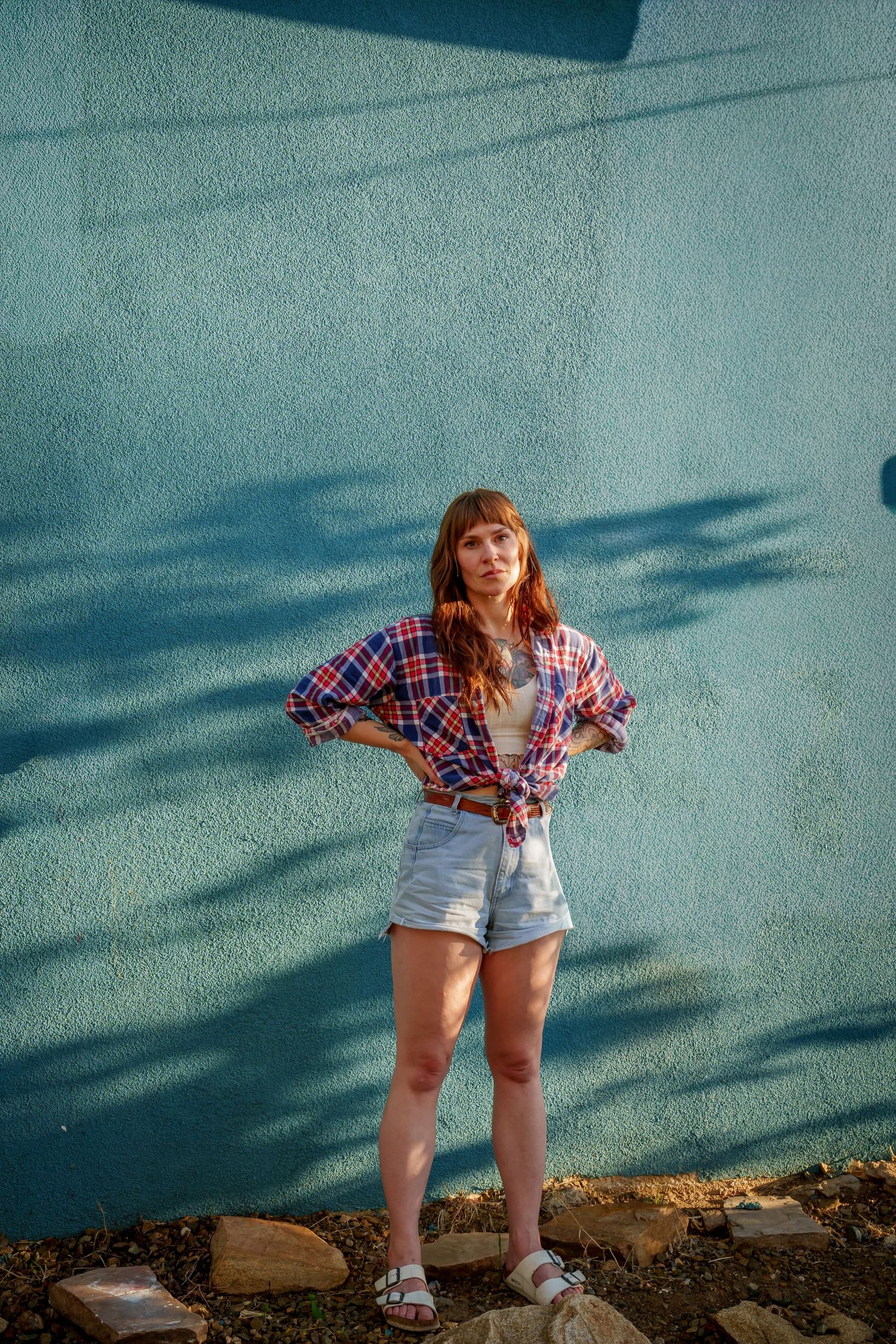 Young woman in plaid shirt and white denim shorts stands confidently against a blue textured wall in sunlight.