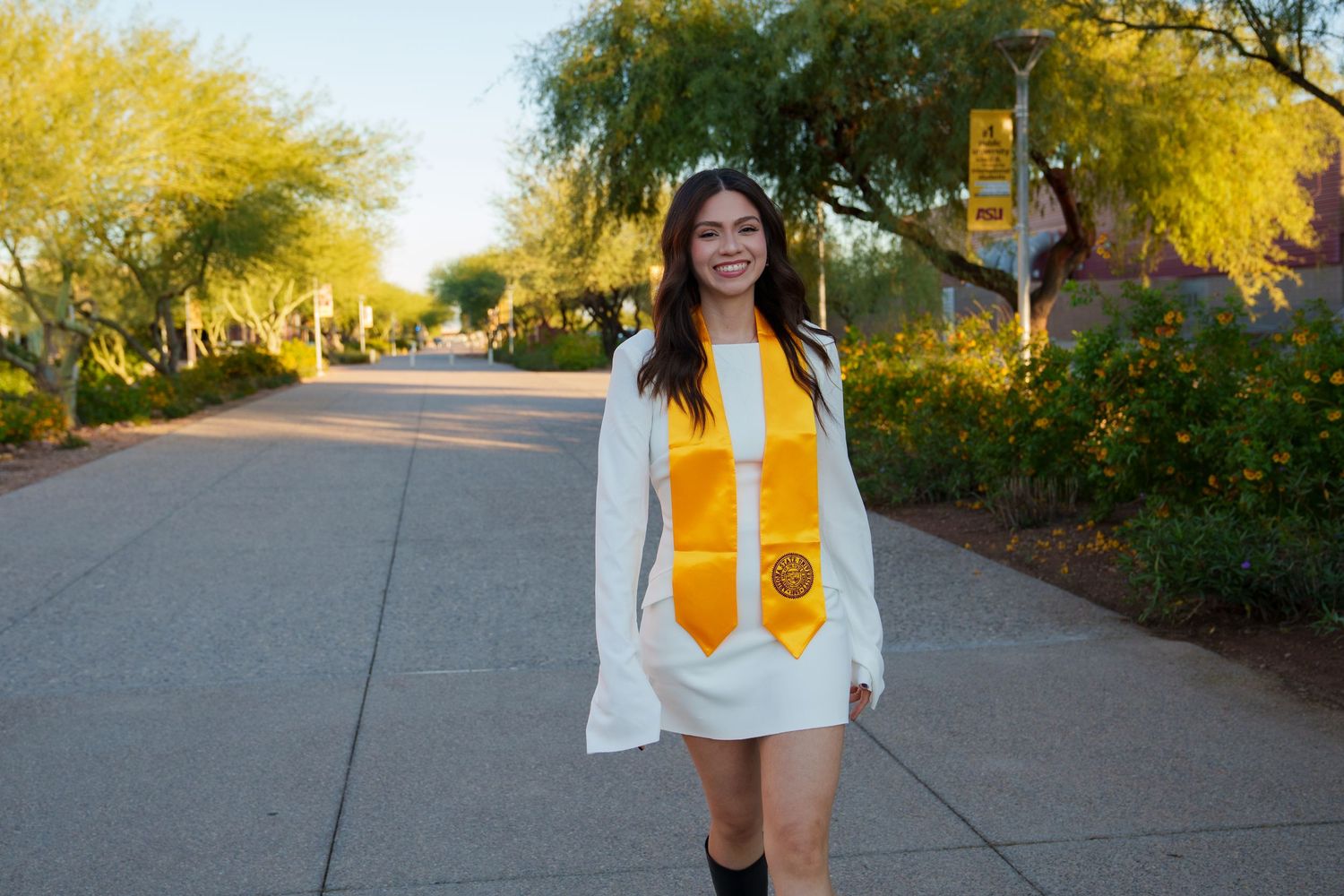 College graduate smiles in white graduation gown and gold stole walking on a sunny tree-lined campus path.