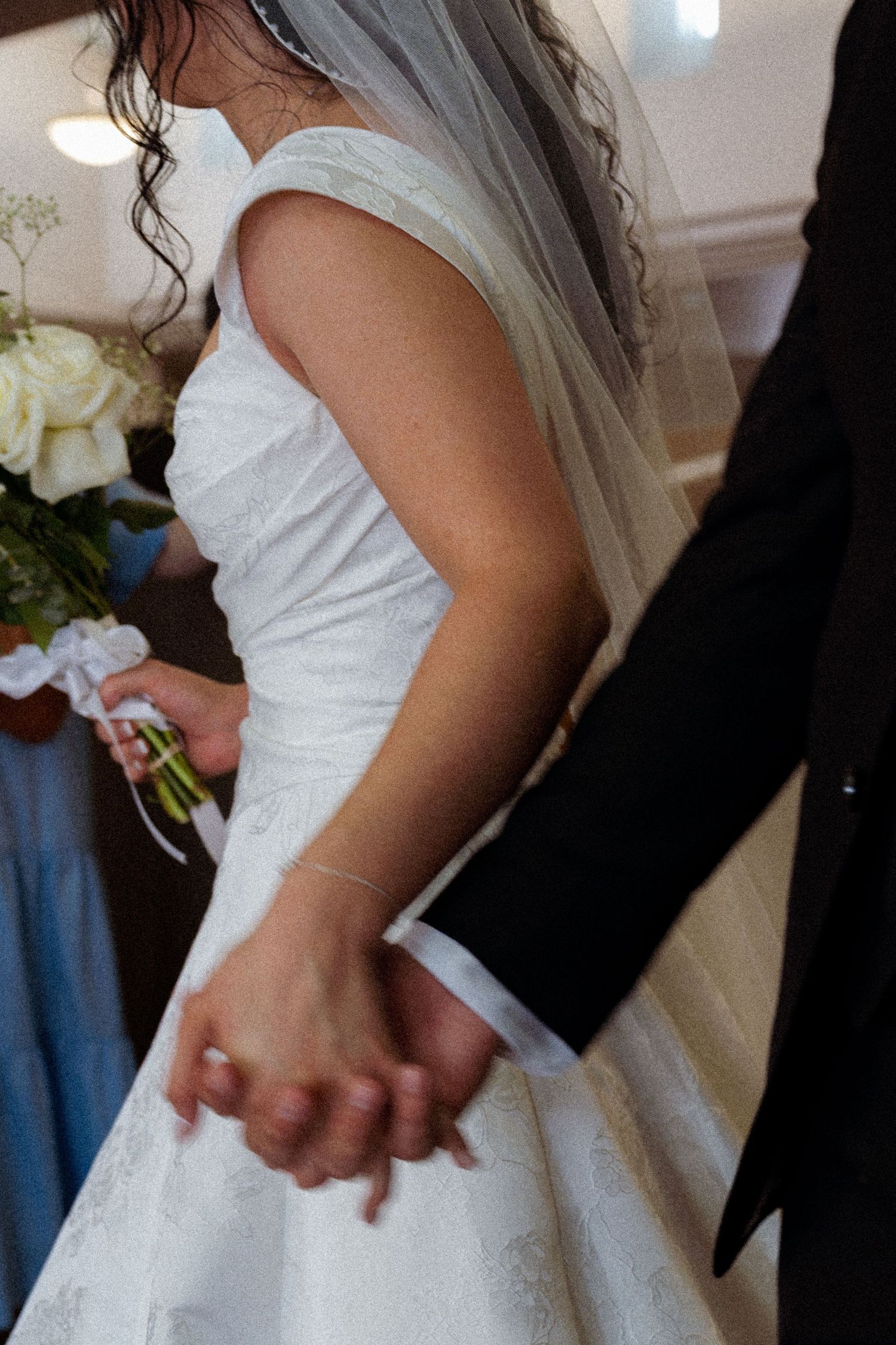 Bride in white lace gown and veil holds hands with groom in black suit during wedding ceremony.
