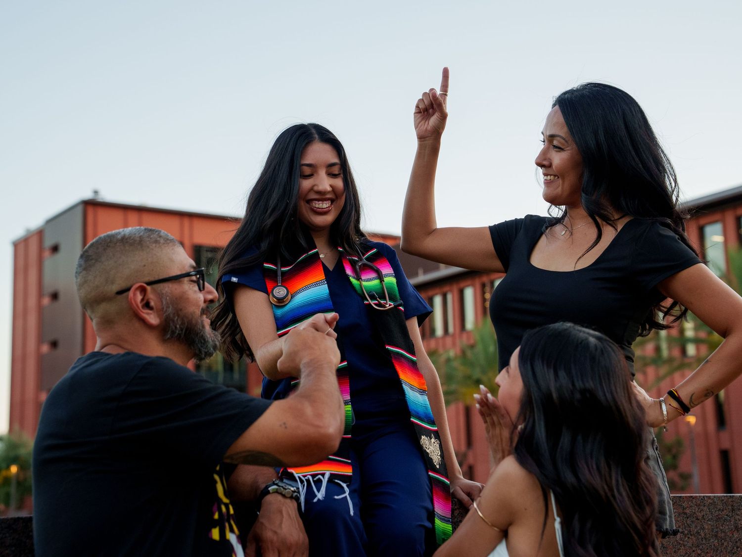 A group of four friends laugh and dance together outdoors at dusk near a brick building.