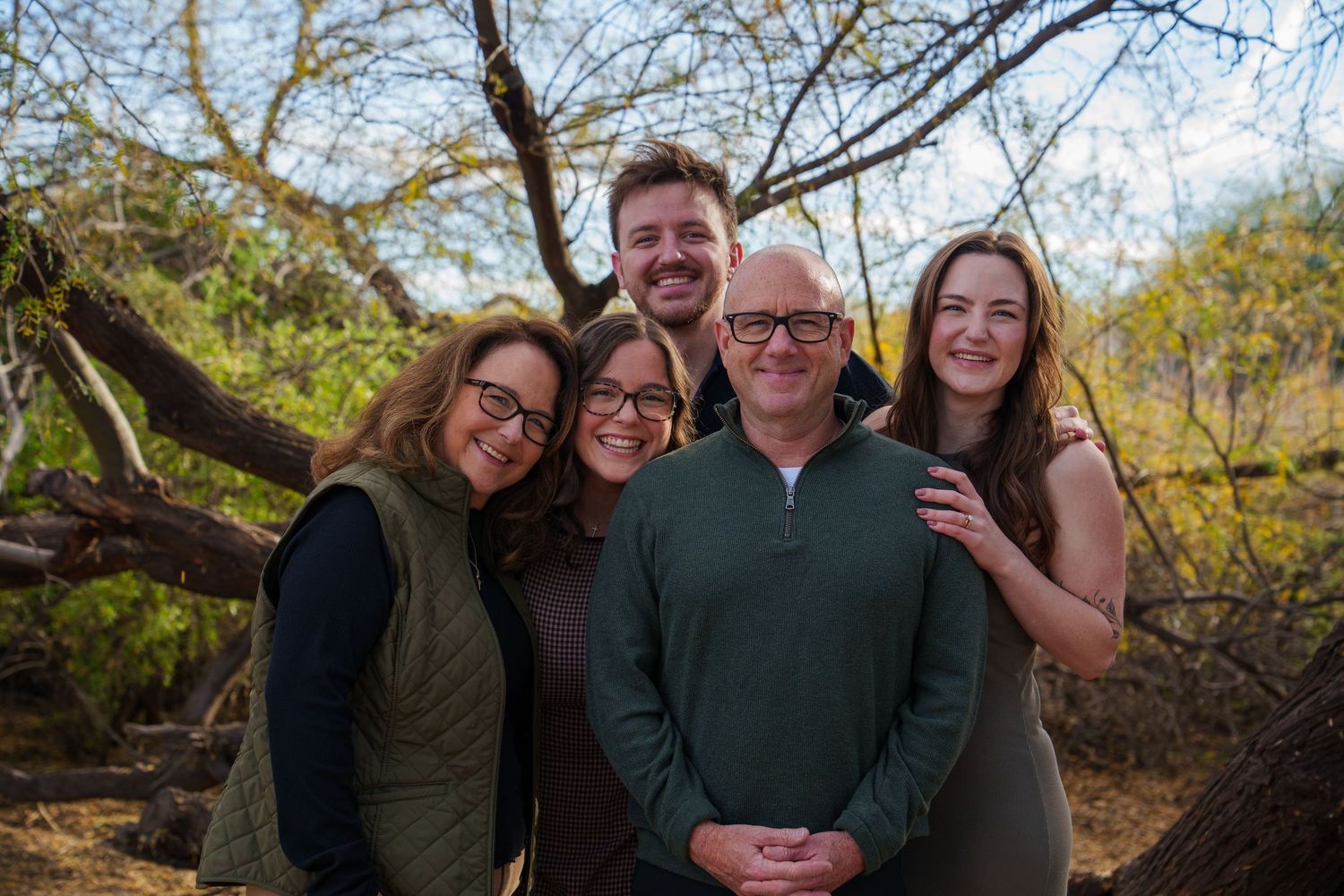 Happy family of five smiling together outdoors in a natural wooded setting during a portrait session.