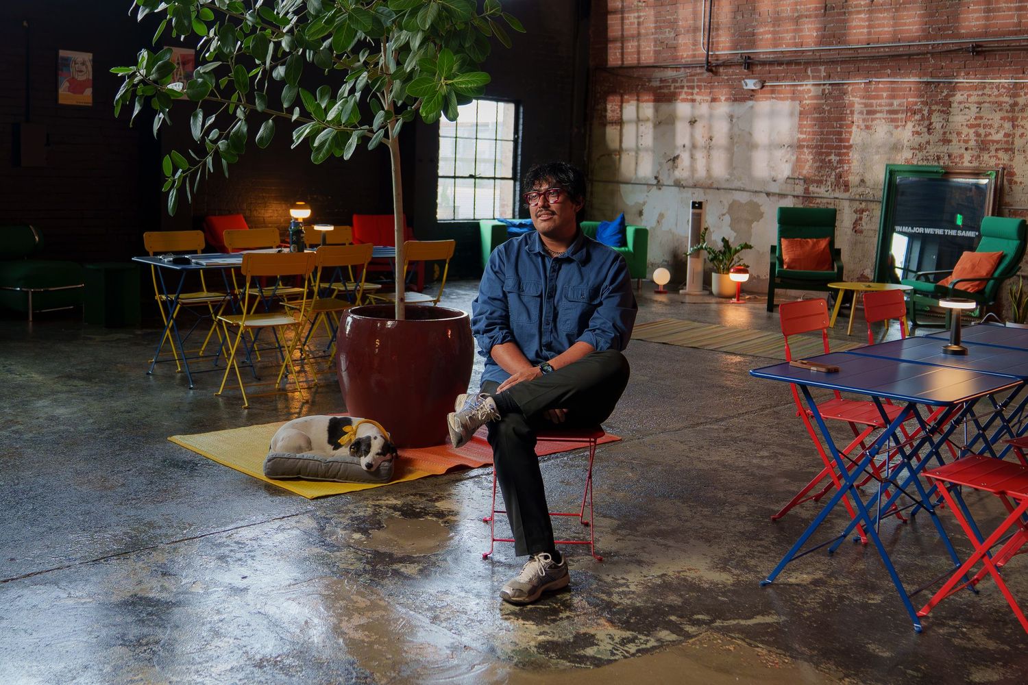 Man sitting on red chair in colorful industrial space with plants, tables, and chairs on concrete floor.