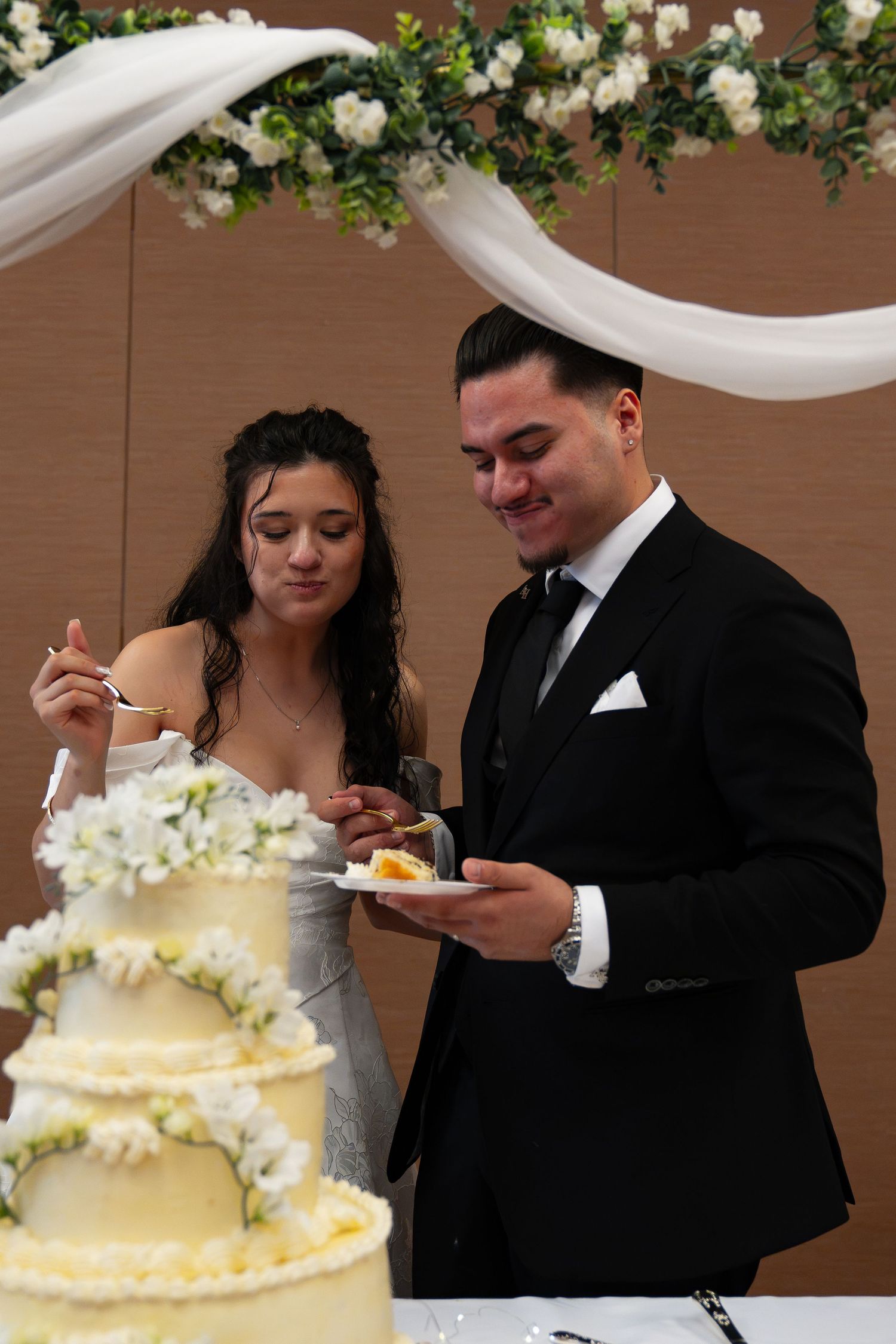 Bride and groom smiling while cutting their white floral wedding cake at elegant indoor reception.