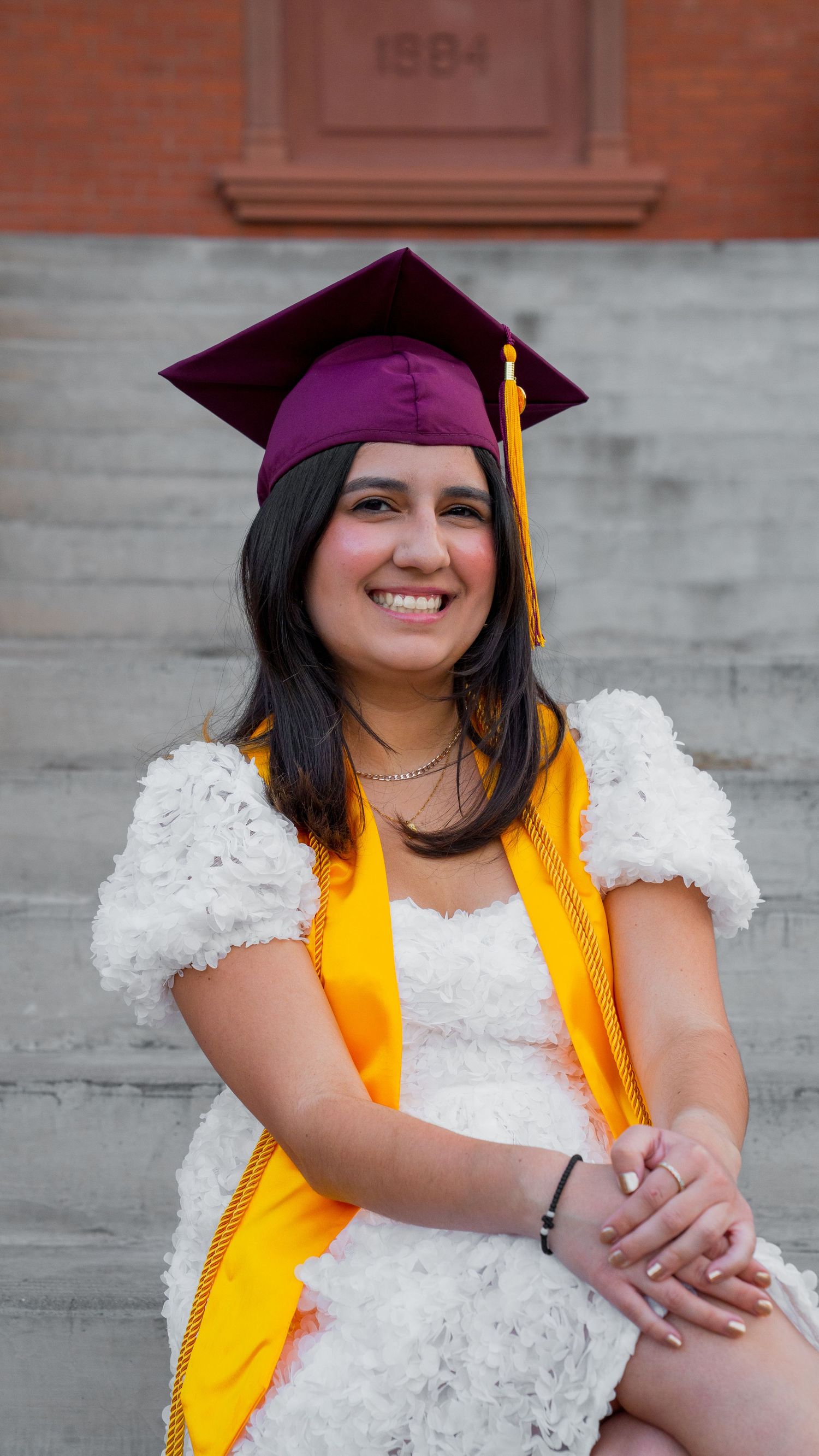 Graduate wearing white lace dress and maroon cap with gold tassel poses on concrete steps near brick wall.