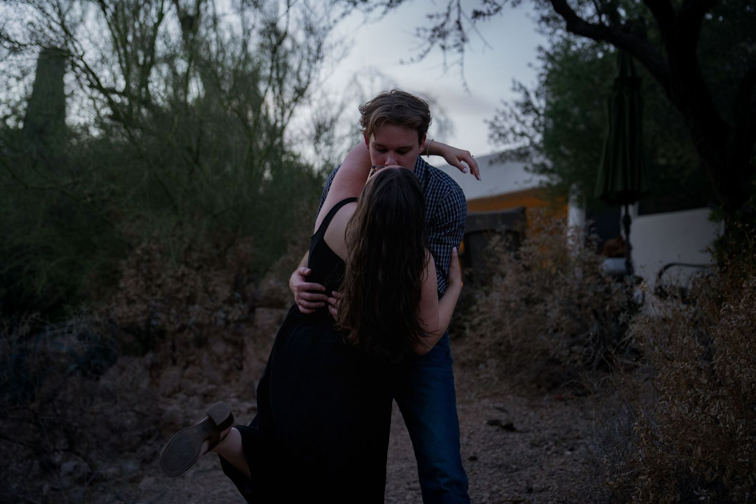 Couple embracing outdoors at dusk near desert trees and a white building in a romantic moment.