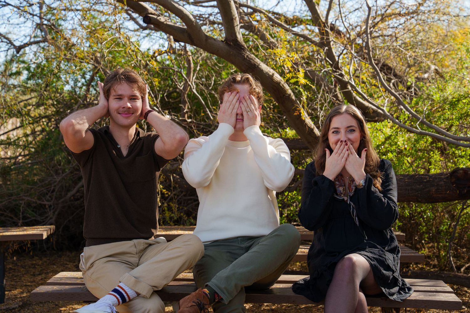 Three friends sitting outdoors posing as the three wise monkeys, see no evil, hear no evil, speak no evil.