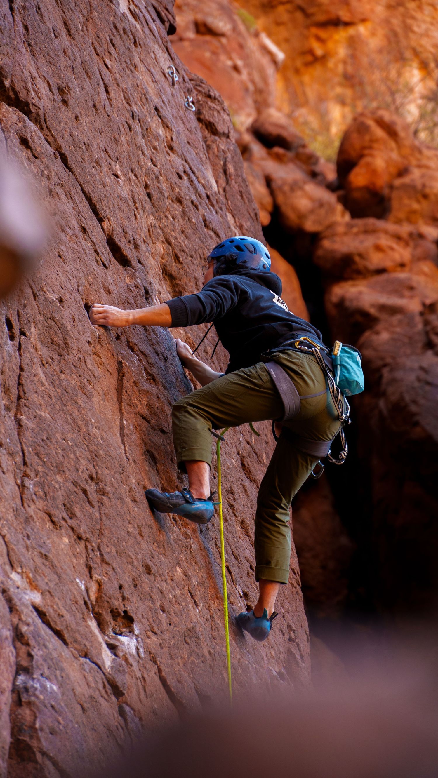 Rock climber traversing horizontally across a red sandstone cliff face with climbing gear.