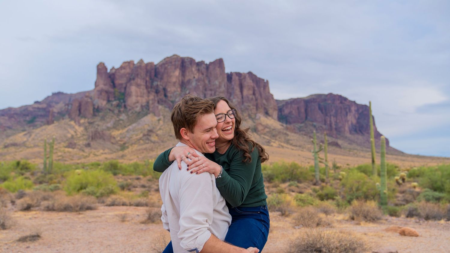 A couple shares an intimate moment against the dramatic backdrop of Arizona's Superstition Mountains at sunset.