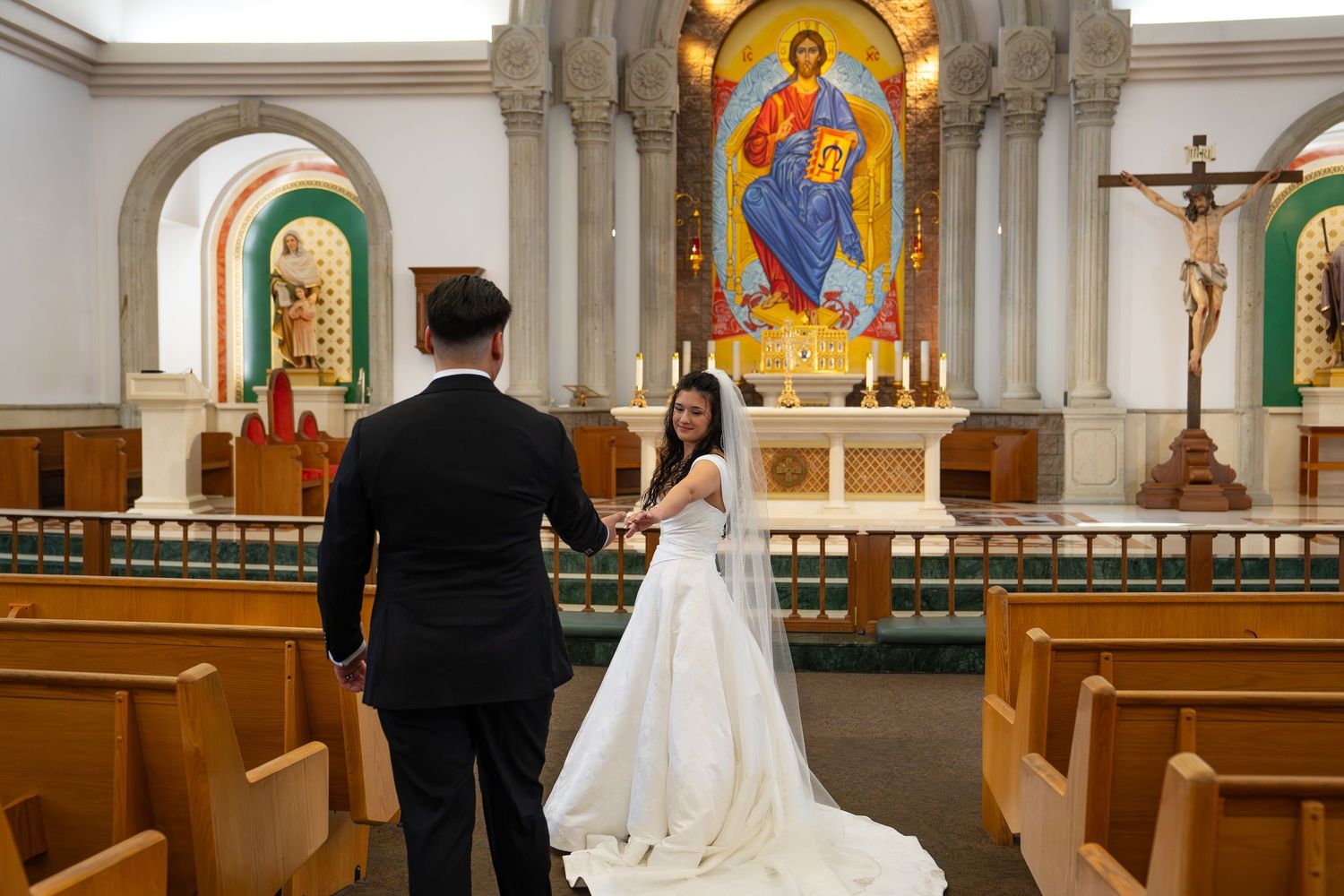 Bride and groom holding hands at church altar with colorful mosaic of Jesus Christ during wedding ceremony.