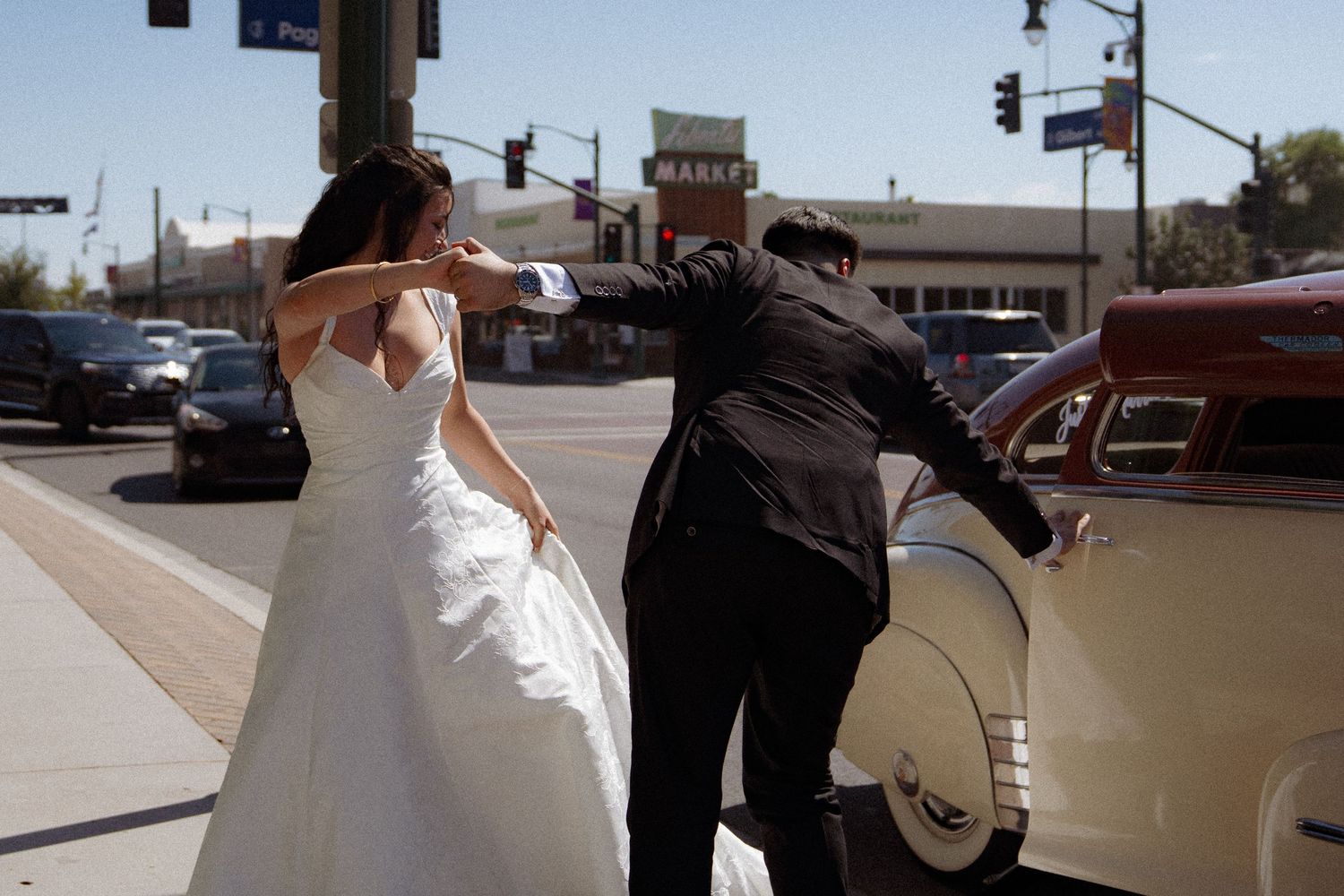 Bride in white gown and groom in black suit pose near vintage car at sunny street intersection.