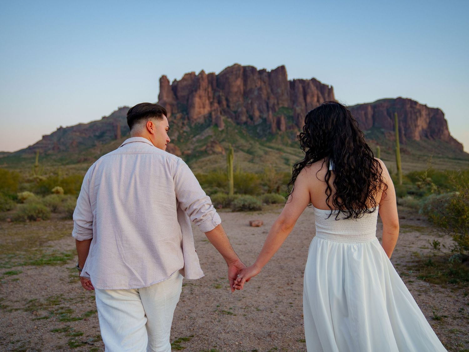 A couple in white outfits holds hands while walking through the scenic Superstition Mountains desert landscape at sunset.