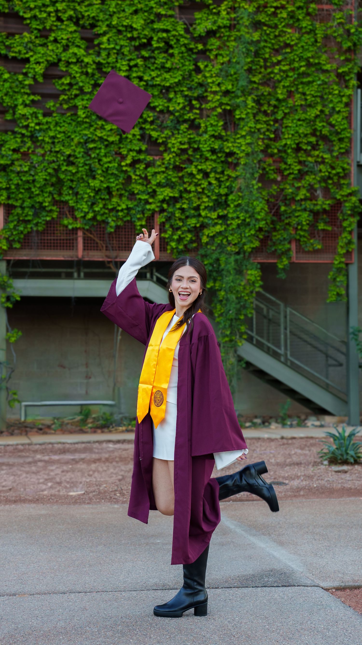 Graduate in purple cap and gown with yellow stole celebrates joyfully on campus grounds with ivy-covered wall backdrop.