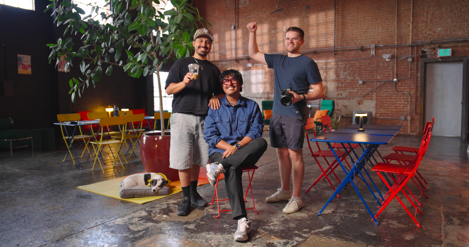 Three young men pose in a colorful industrial space with bistro chairs, plants, and brick walls.