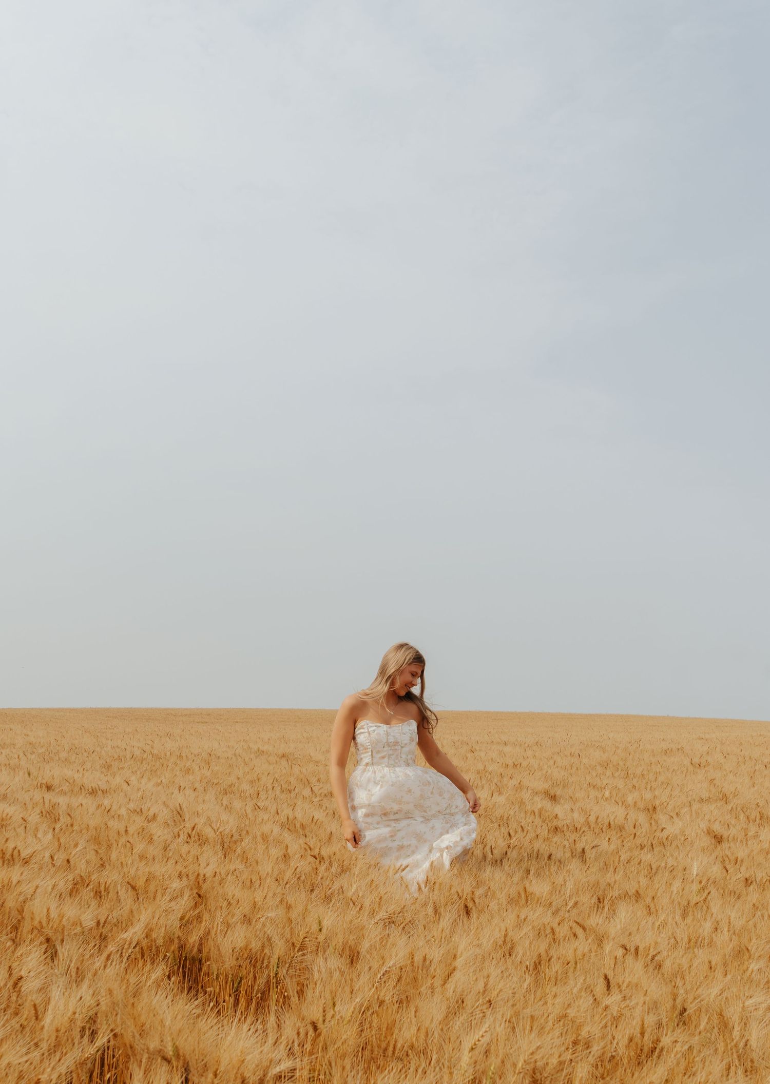 A bride in a white strapless gown walks through a golden wheat field under a soft gray sky.