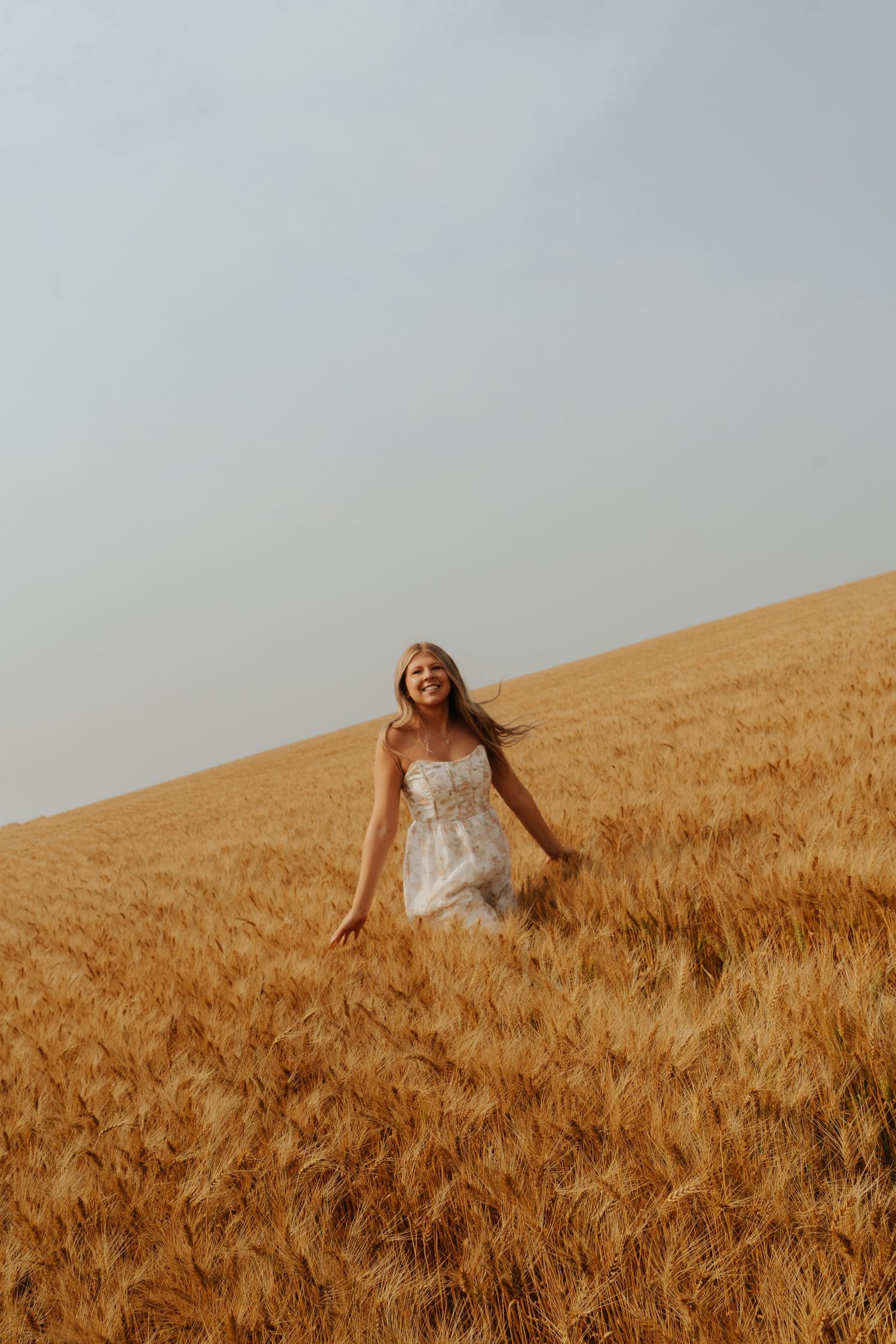 A joyful woman in a white dress walks through a golden wheat field under a soft gray sky.