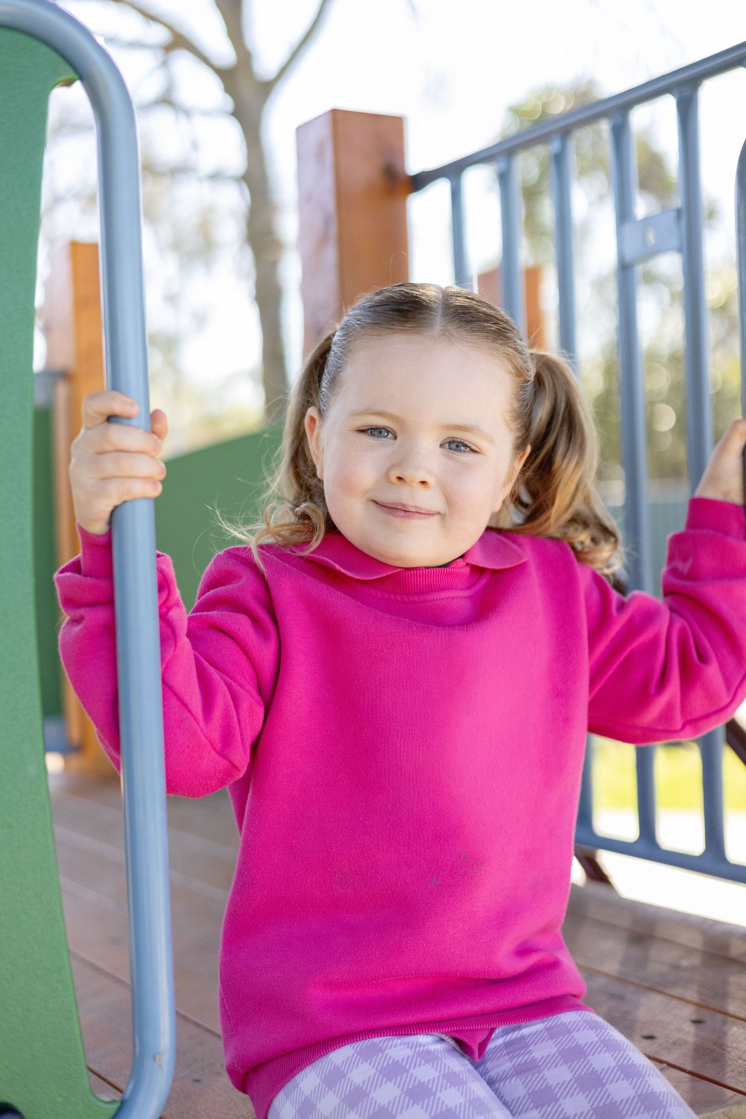 Young girl in pink sweatshirt smiling at playground equipment on sunny day.