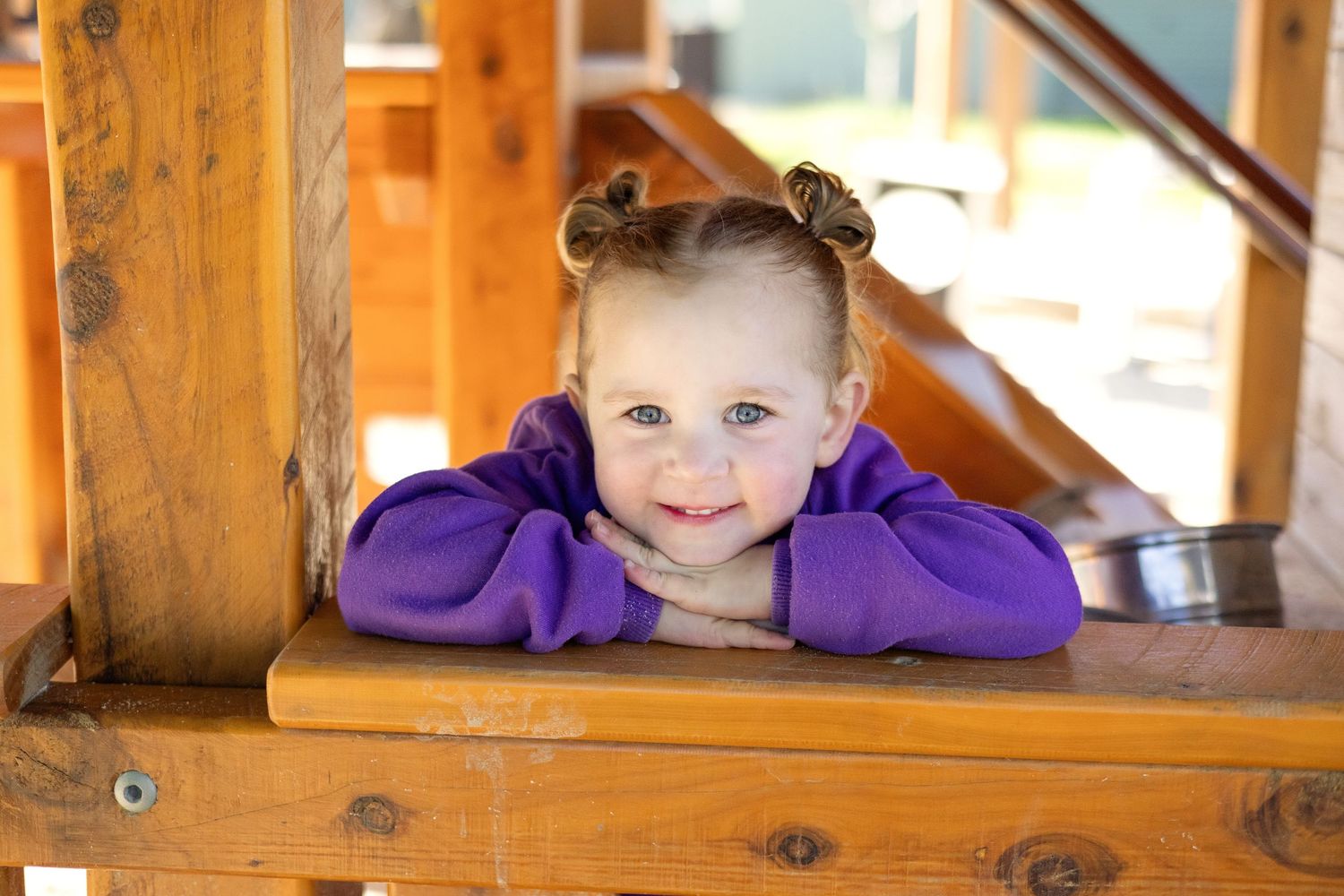 Smiling toddler girl with pigtails resting on wooden playground structure wearing a purple sweater.