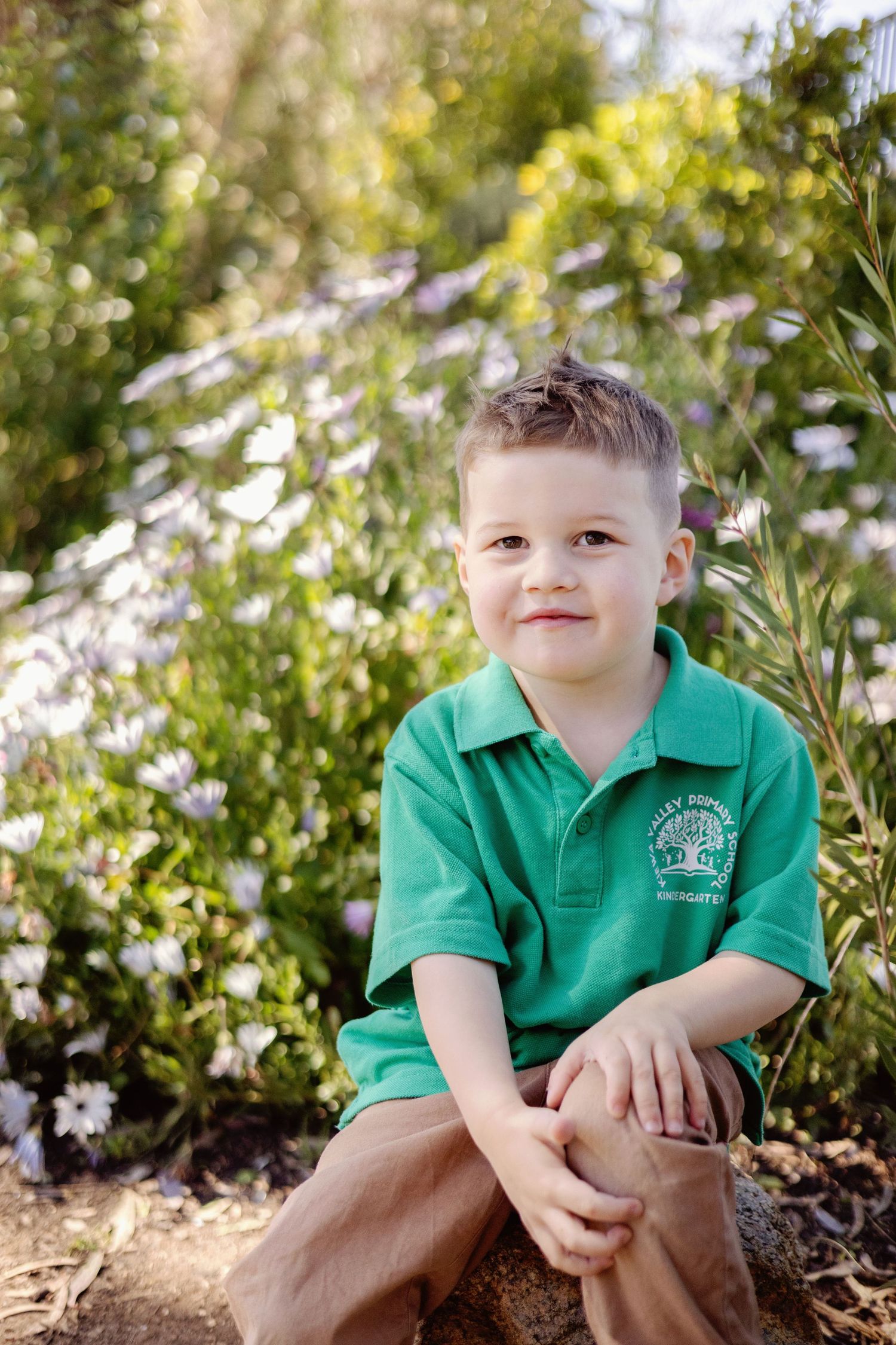 Young boy in green polo shirt sits smiling among wildflowers in a sunny garden.