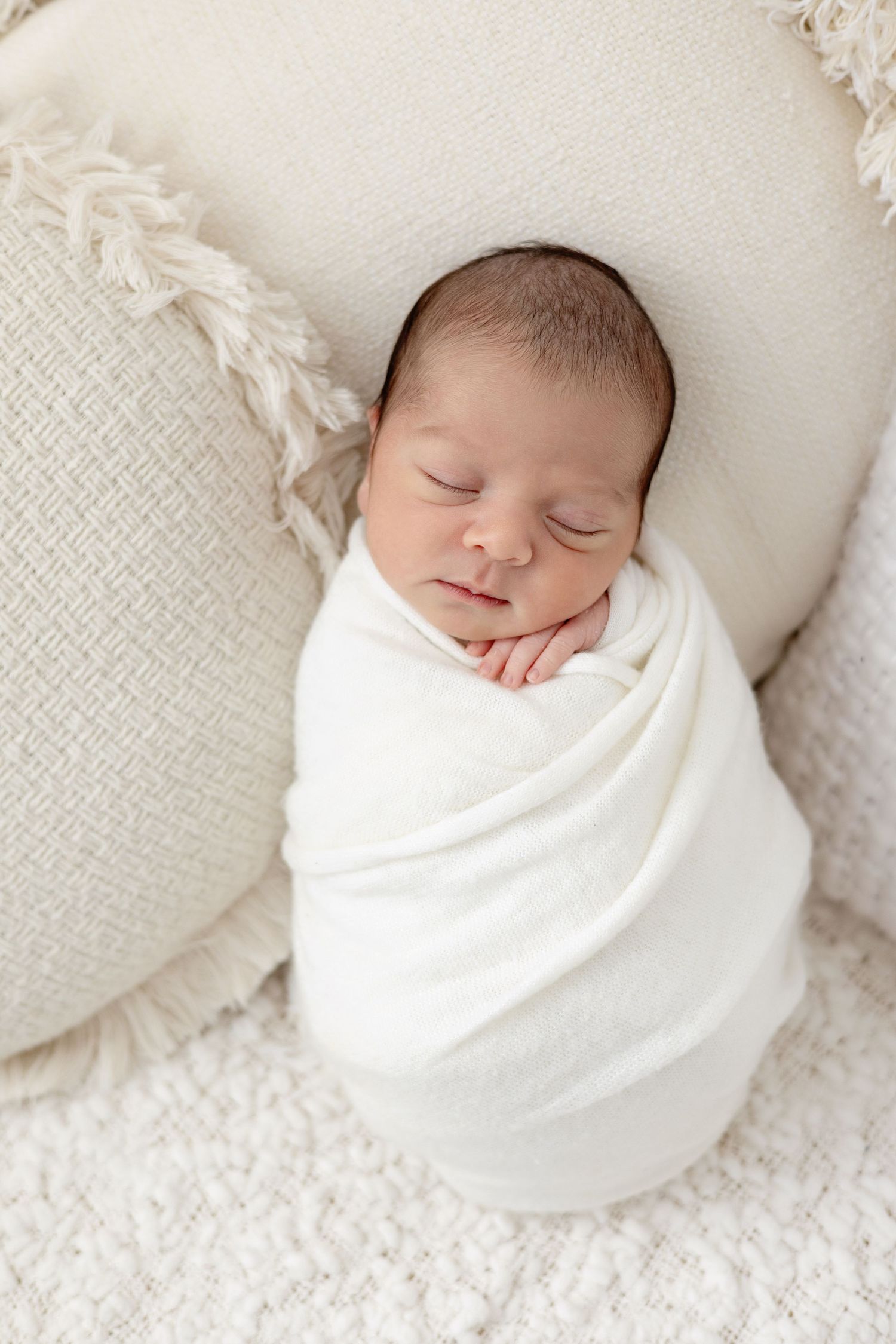 Peacefully sleeping newborn baby swaddled in white wrap, resting on cream textured pillows in a serene studio setting.  Newborn Photography