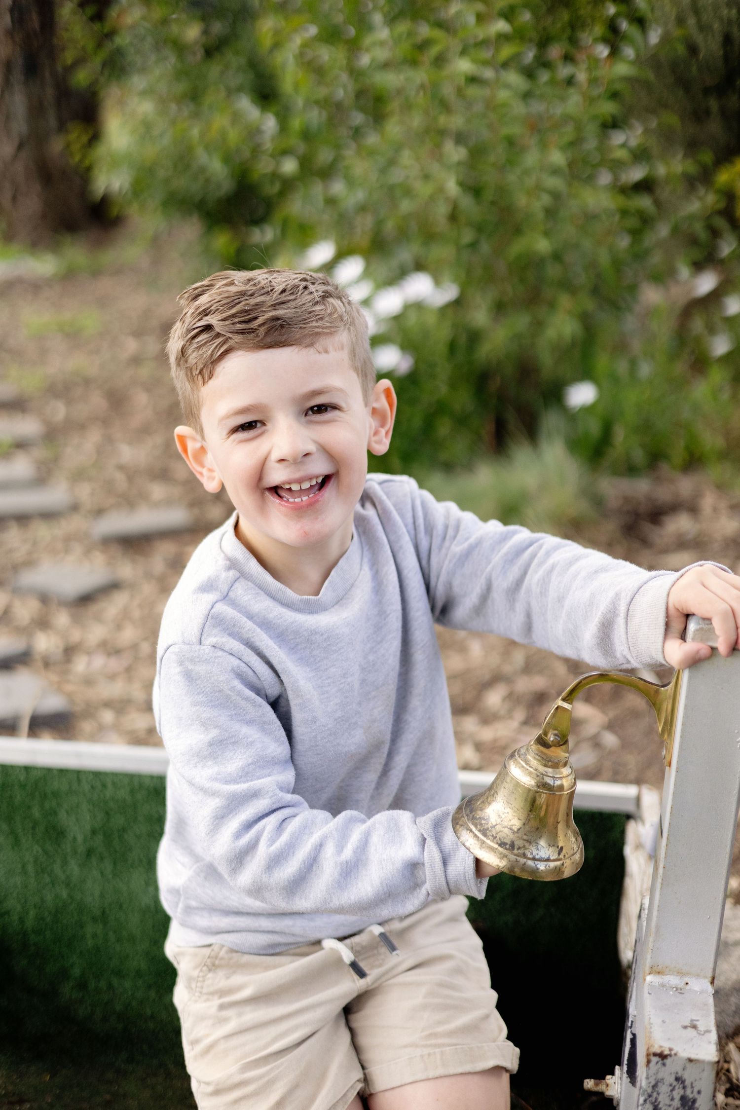 Happy young boy laughing while holding a brass bell outdoors in a garden setting.
