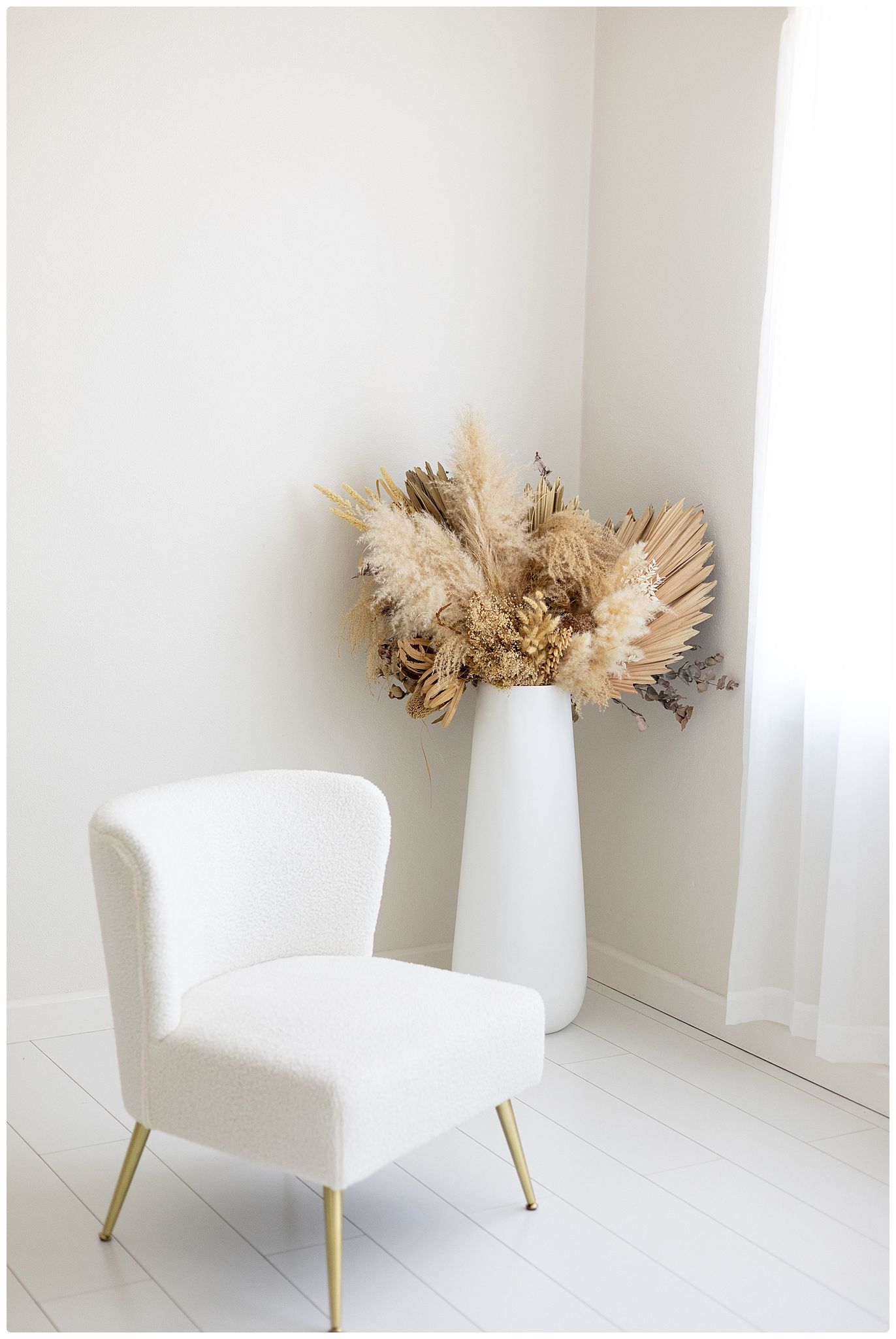 White boucle accent chair with gold legs beside a tall white vase holding dried pampas grass in a bright minimalist corner.