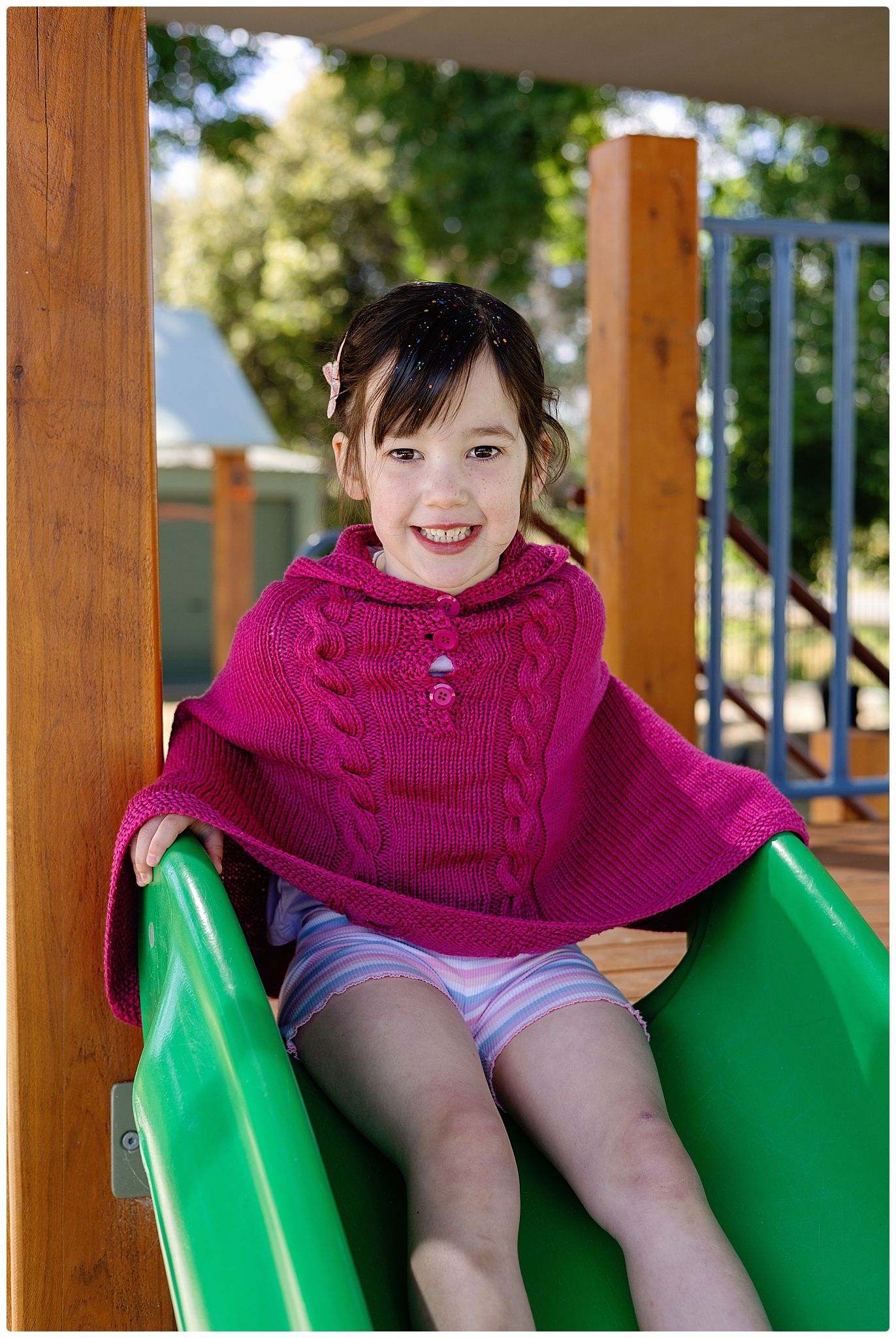 Smiling young girl in pink knit poncho sitting on a green playground slide outdoors.