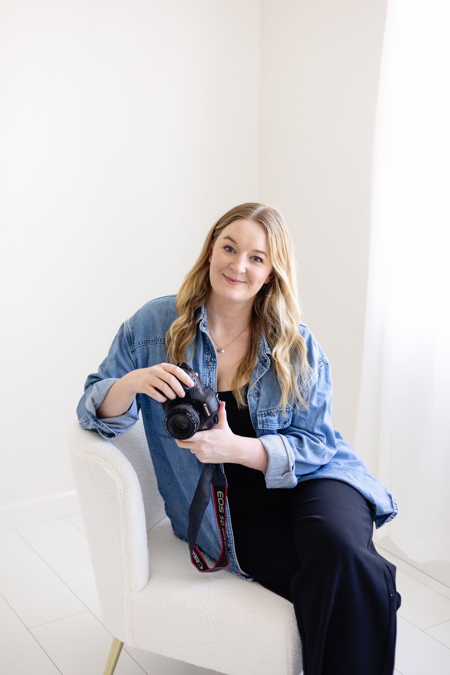 Smiling blonde woman photographer sitting in white chair holding a Canon DSLR camera, wearing denim shirt and black pants.