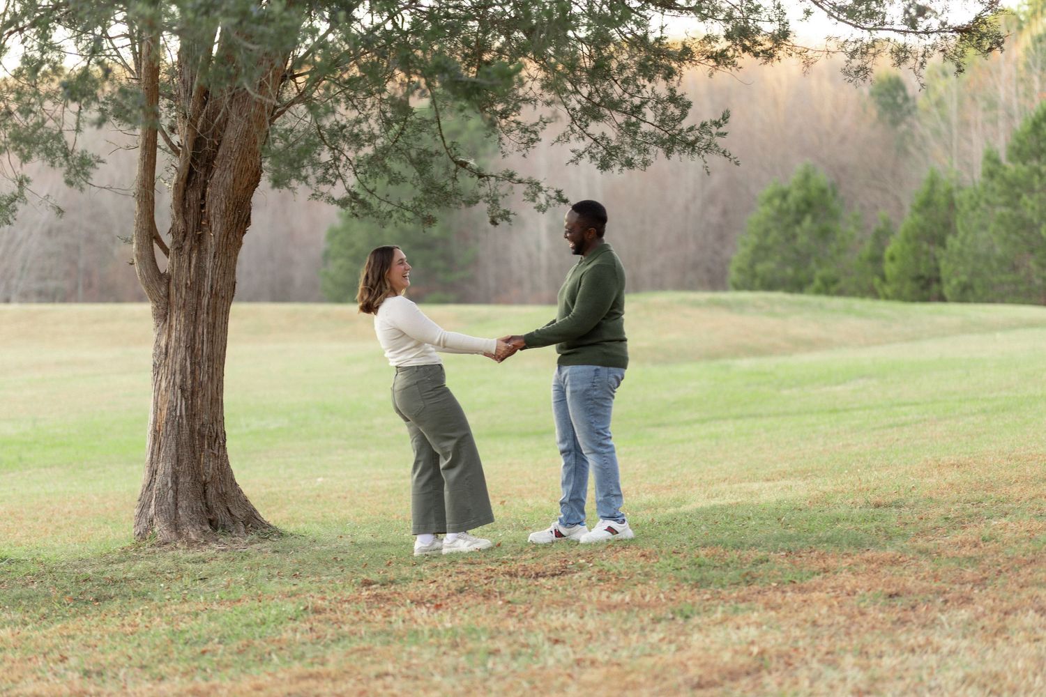 Man and woman laughing and holding hands under a large tree in an open field, enjoying a serene and romantic outdoor moment.