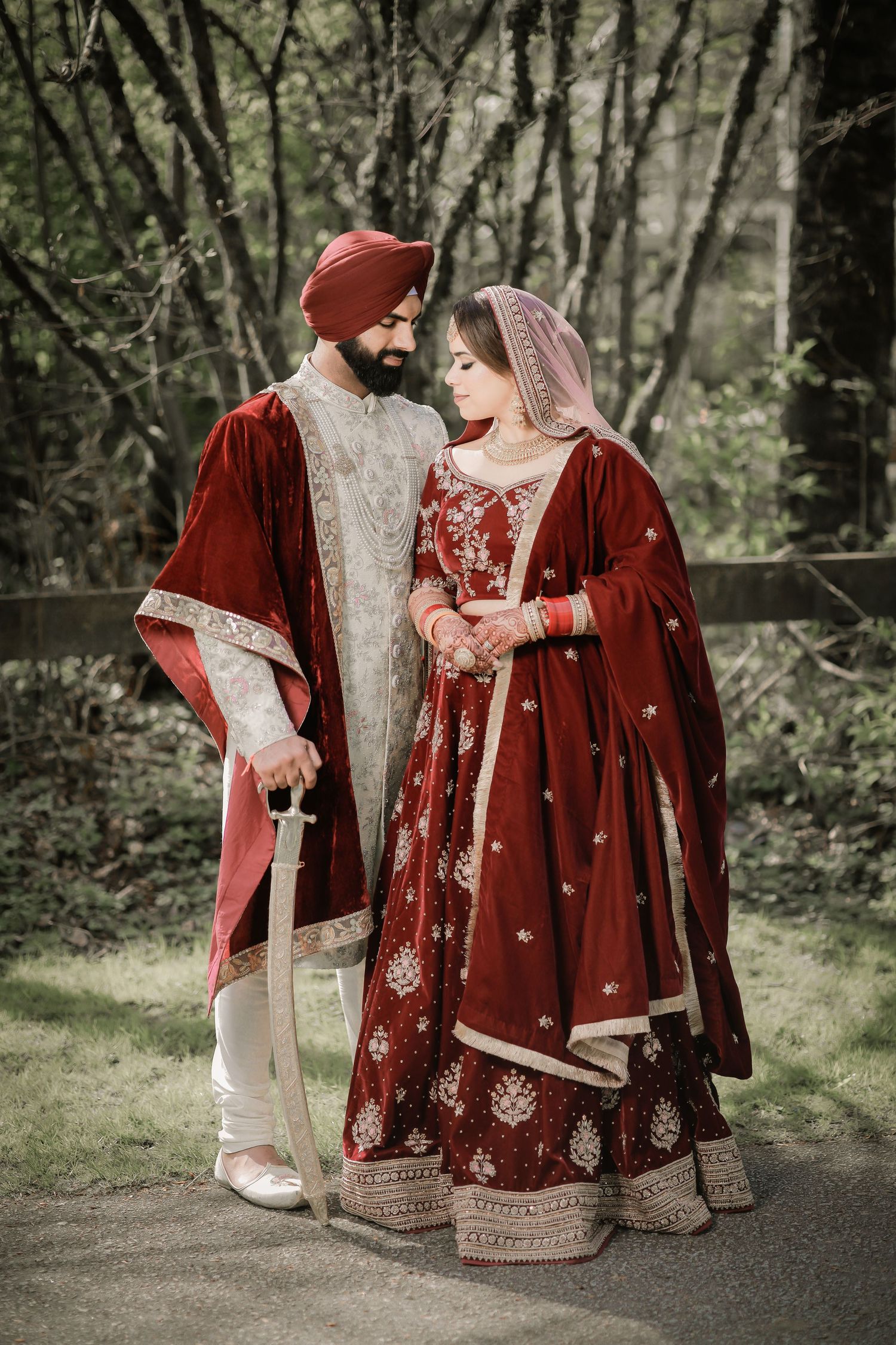 Punjabi bride and groom in red and gold attire, forest backdrop wedding portrait