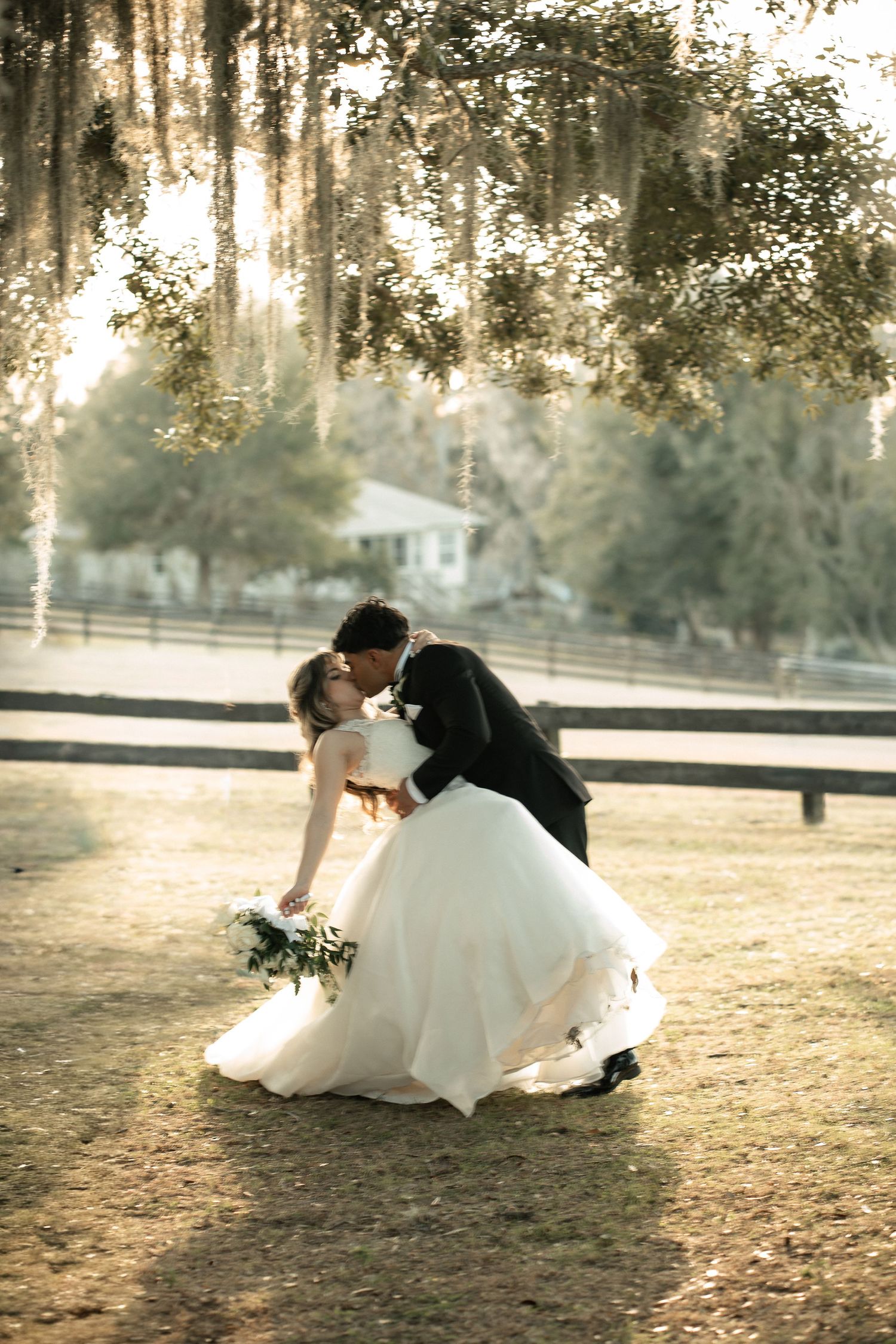 Romantic couple in wedding attire share a tender dip pose at sunset in a rustic outdoor setting with white fencing.