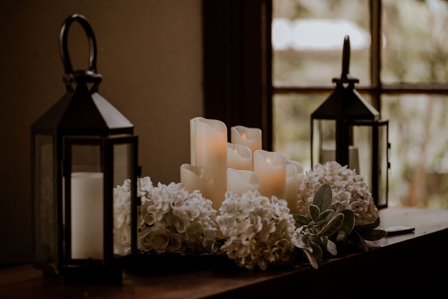 Elegant candle display with white pillar candles, hydrangeas, and black lanterns on a wooden surface near a window.