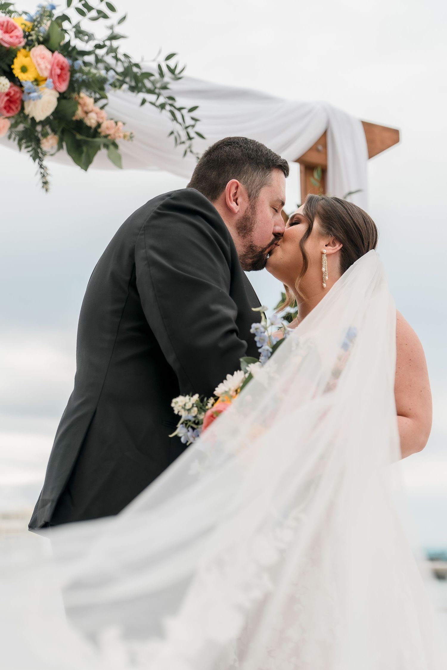 A romantic wedding kiss moment under a floral arch with flowing veil.