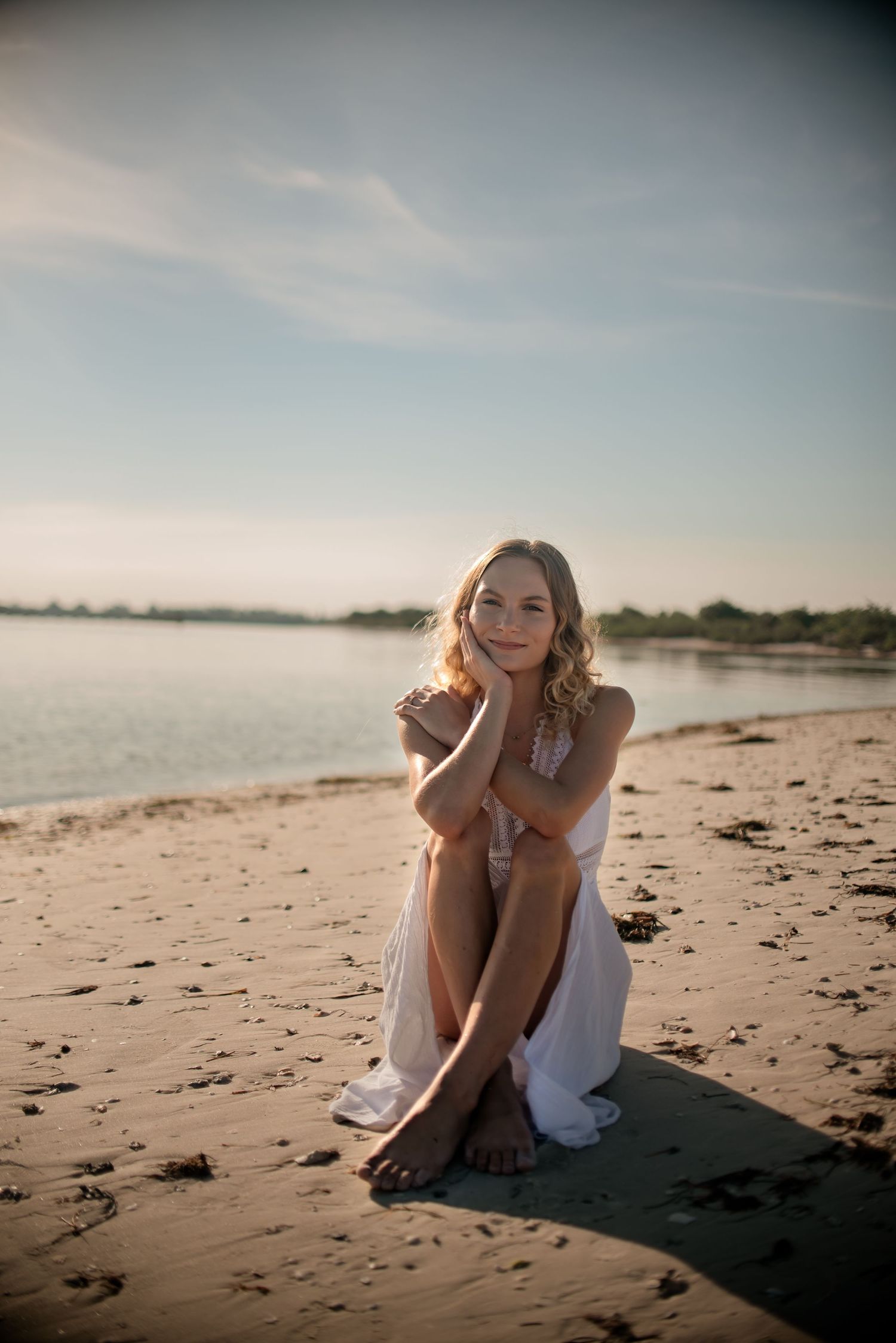 Sunset beach portrait of woman in white dress sitting on sandy shore.