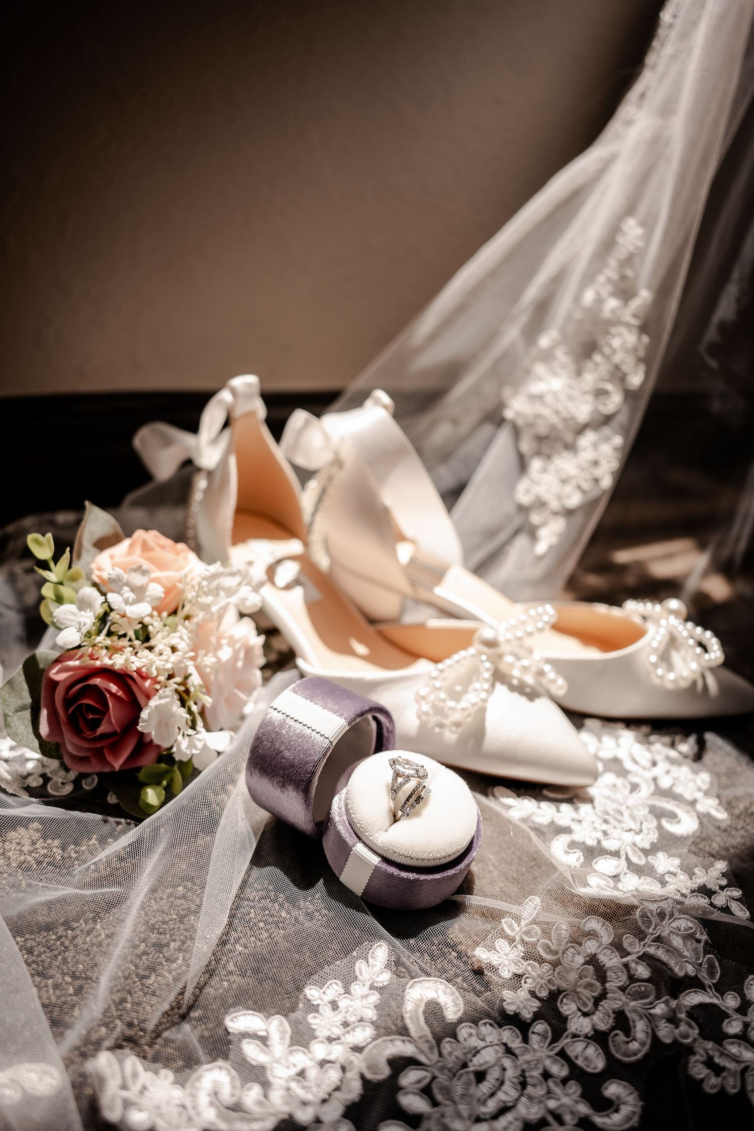 Bridal flat-lay of white satin bow shoes, an engagement ring in a purple box, a rose bouquet and lace veil on a dark surface.