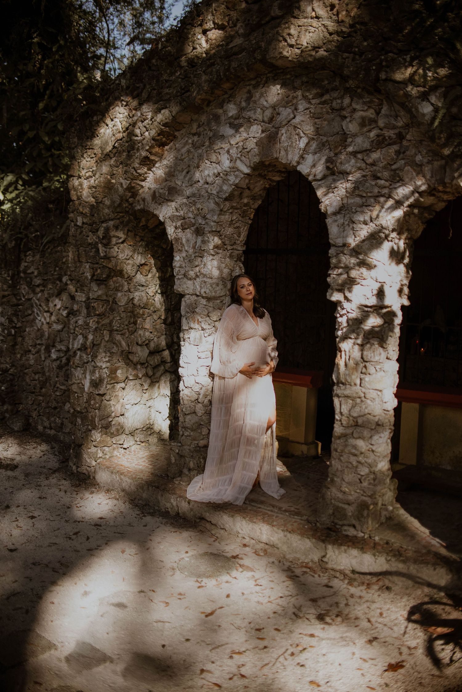 Pregnant woman in white sheer gown standing in sunlit stone archway, cradling baby bump during maternity photo session.