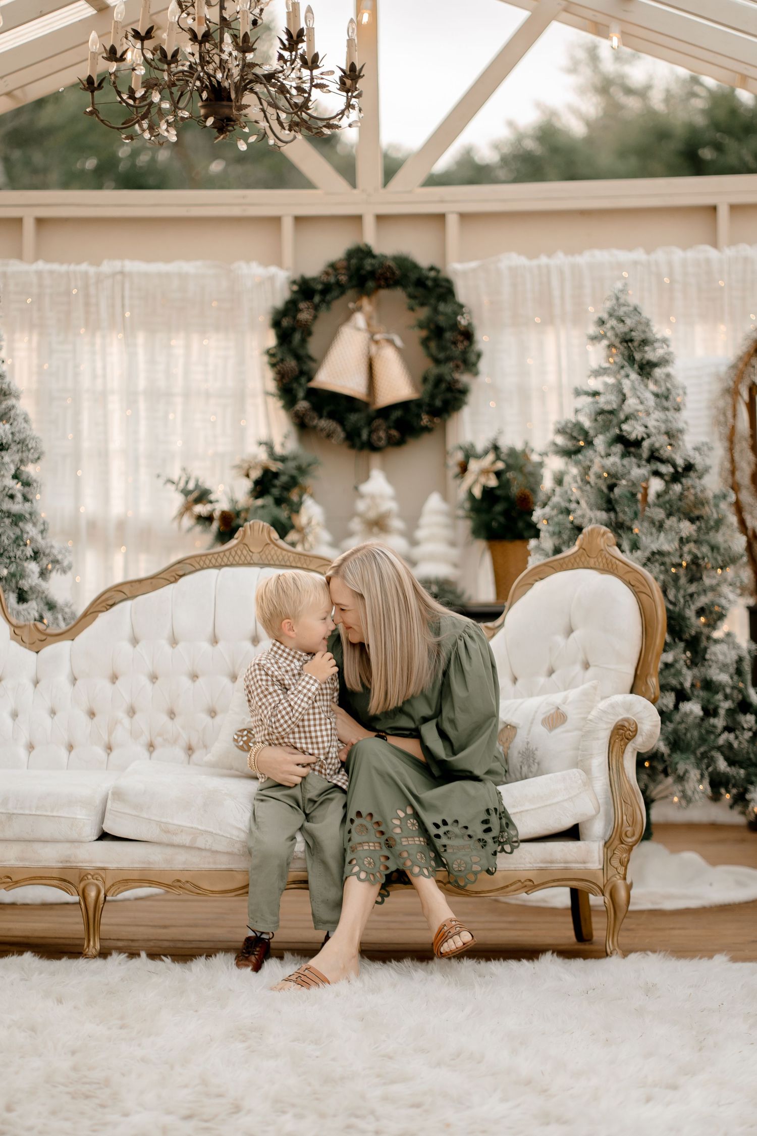 A cozy Christmas scene with people on an elegant sofa surrounded by decorated trees and a wreath in a bright sunroom.