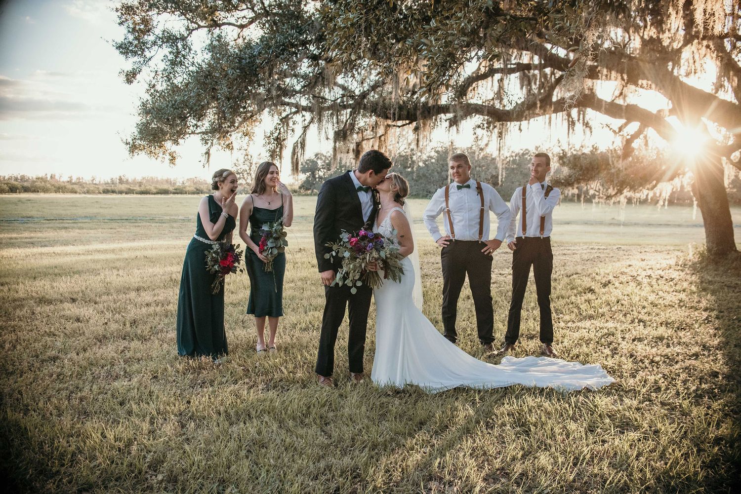 Wedding party poses under a majestic oak tree at sunset in an open field.
