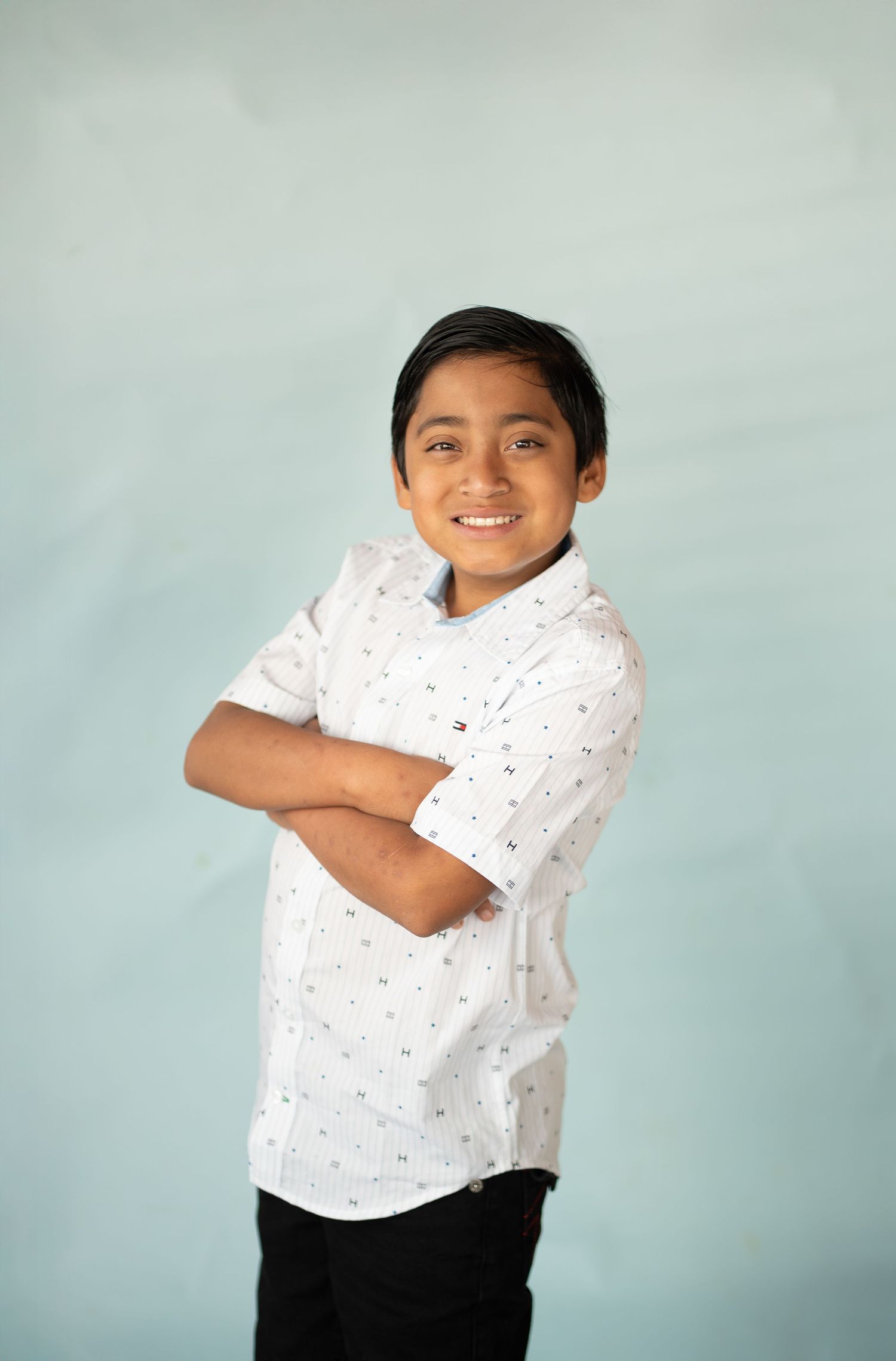 Smiling boy in white patterned shirt with arms crossed poses against light blue background.