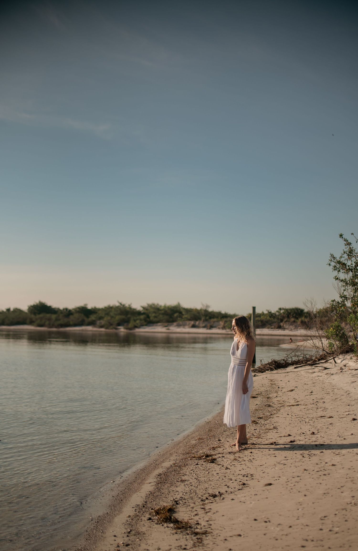 A person in white strolls down a peaceful sandy beach beside calm waters.